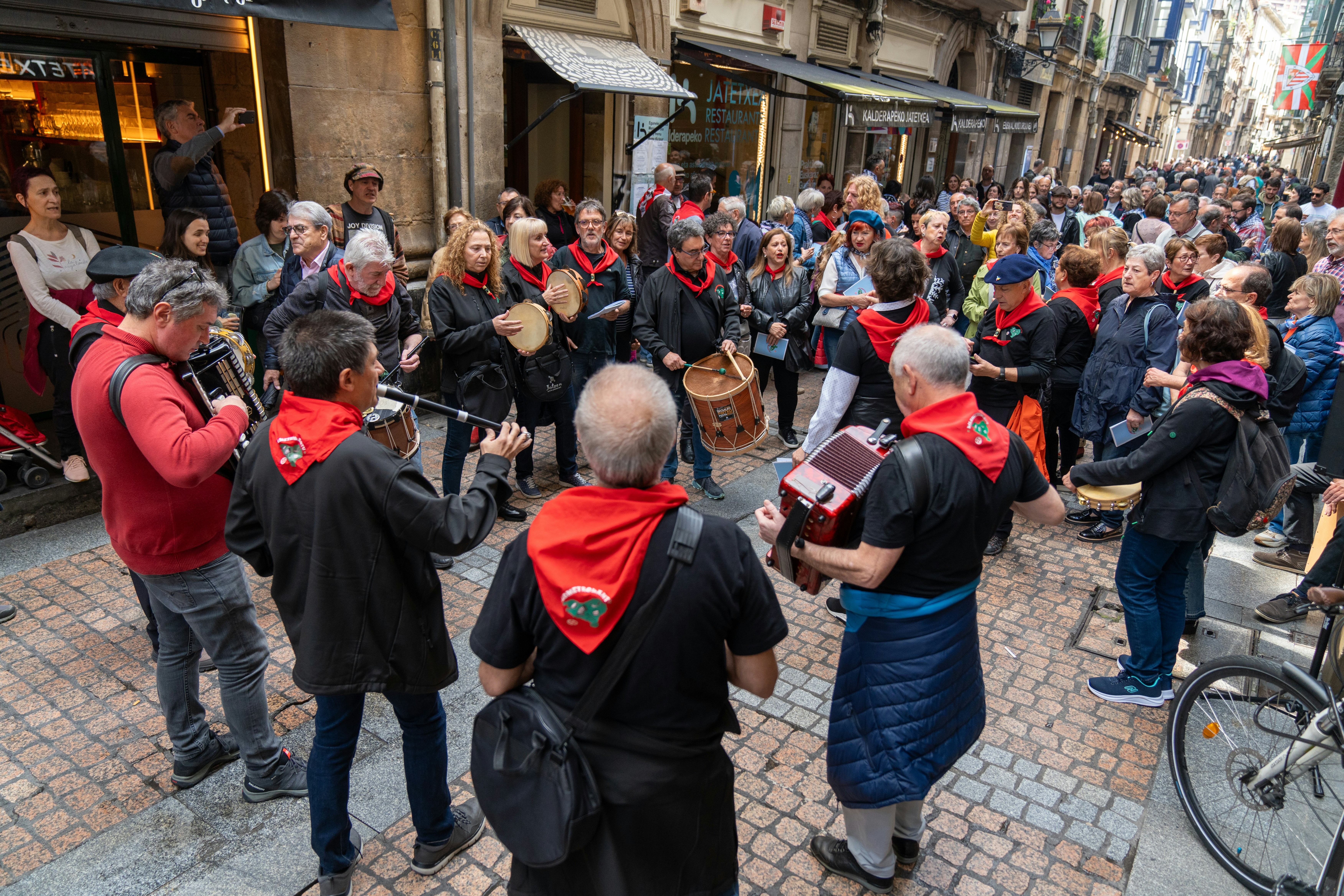 People in red kerchiefs play instruments for a crowd in a narrow city street.