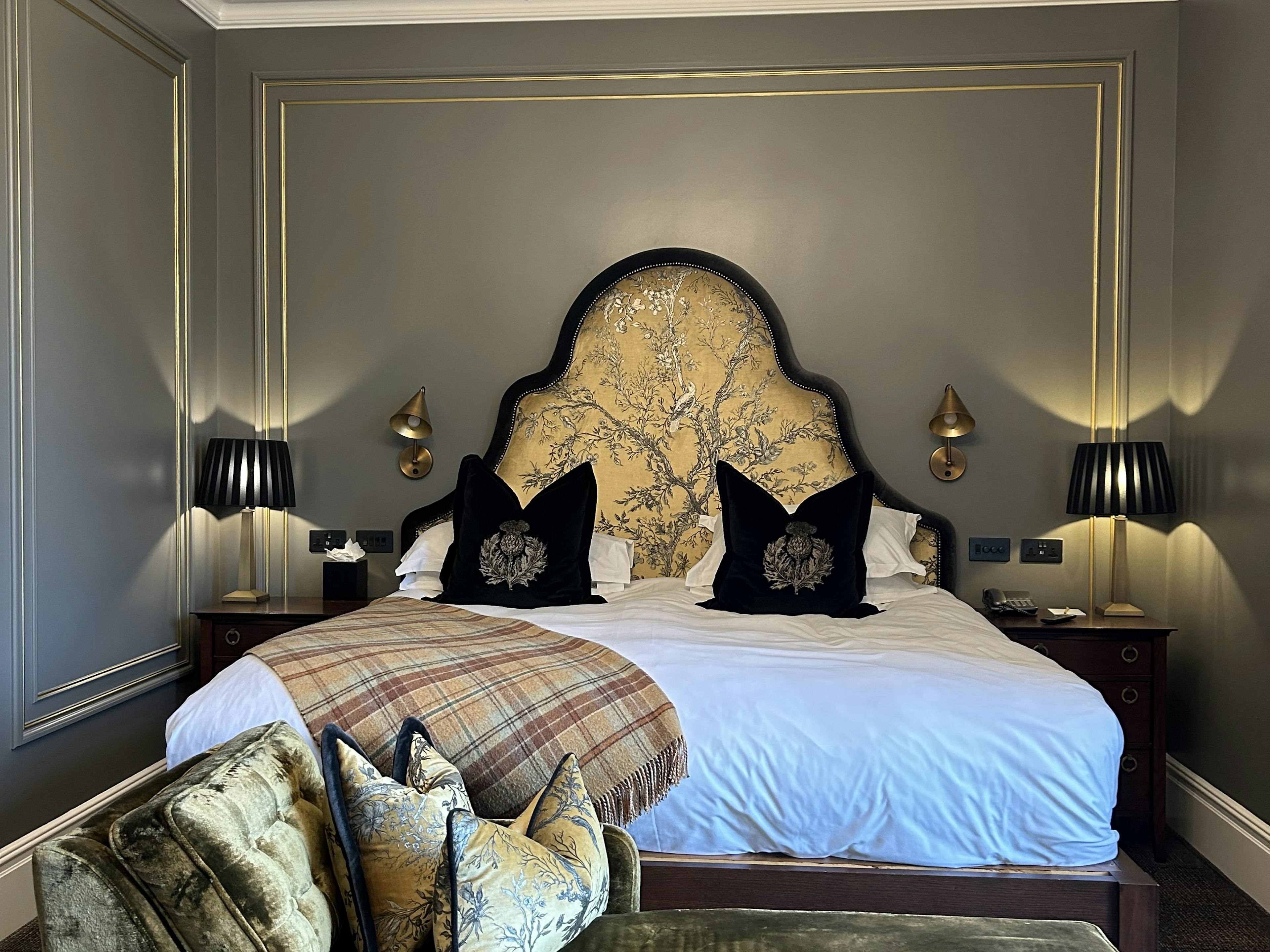 A bed with a white covering has a plaid blanket laid on top and decorative black pillows. The triangular headboard is golden fabric, the bedside tables have lamps, and the walls are gray.