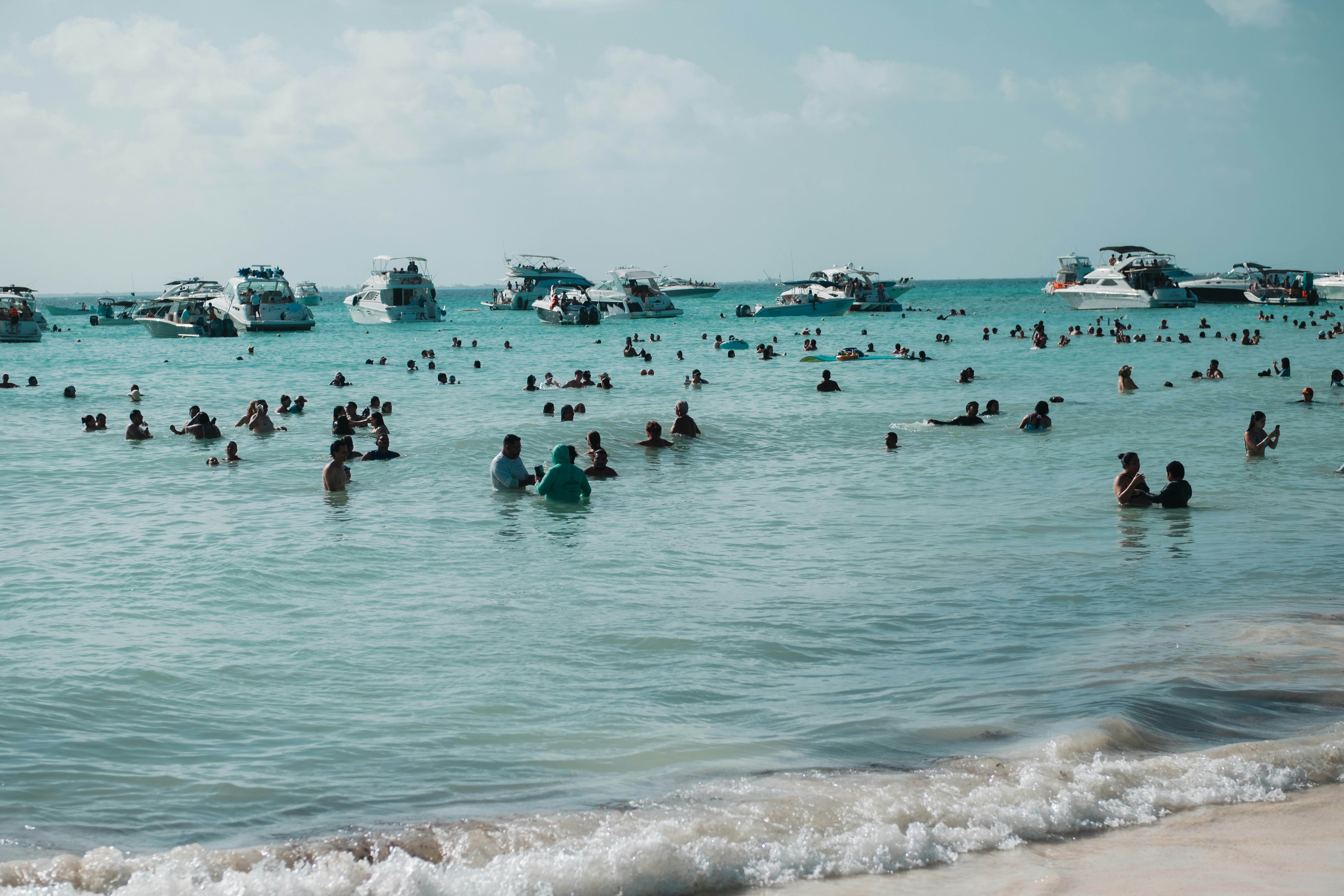 People stand and swim in blue water just off a sandy beach; several boats are stopped offshore.