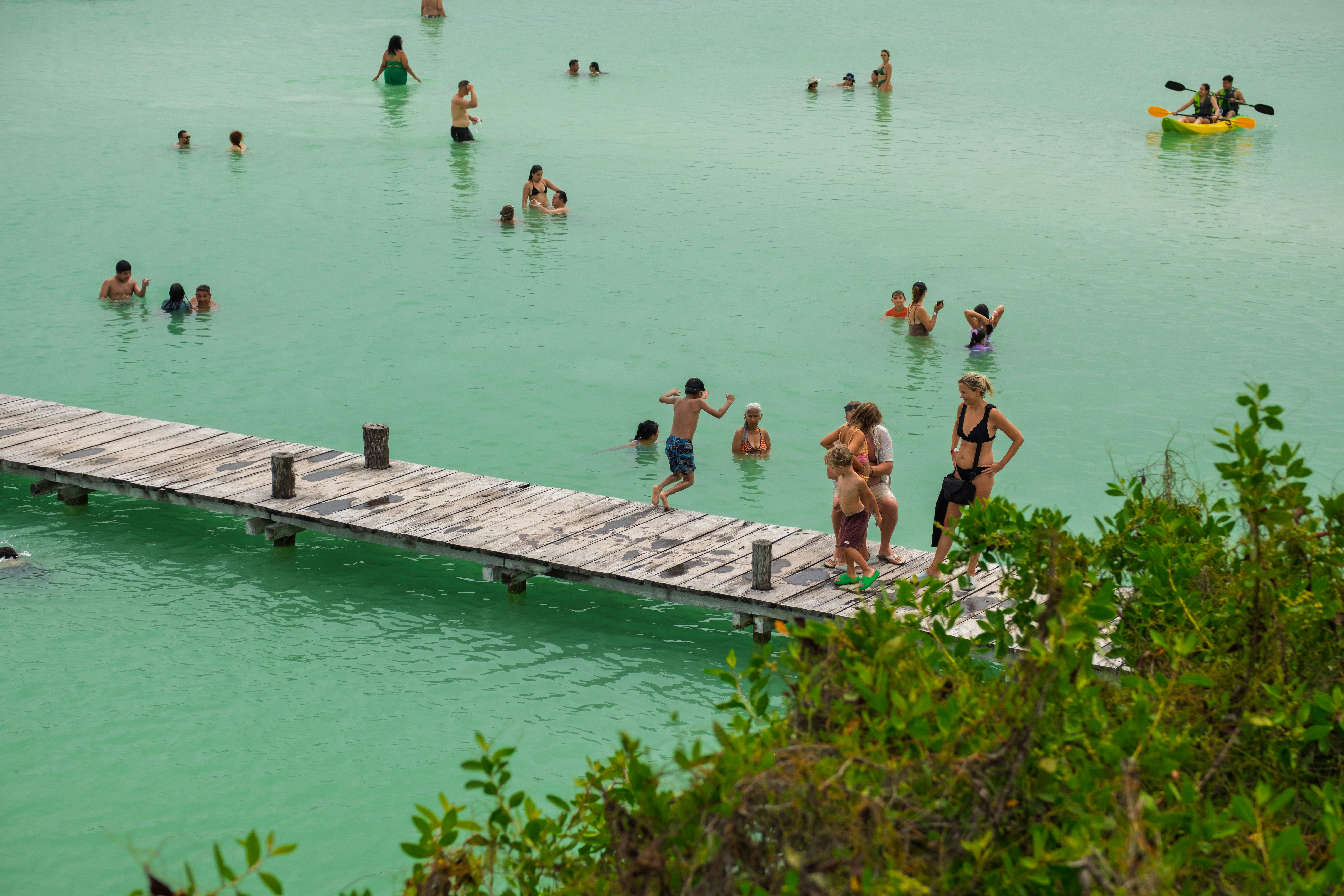 A person jumps from a long wooden pier into a greenish lake where people are standing in the water.
