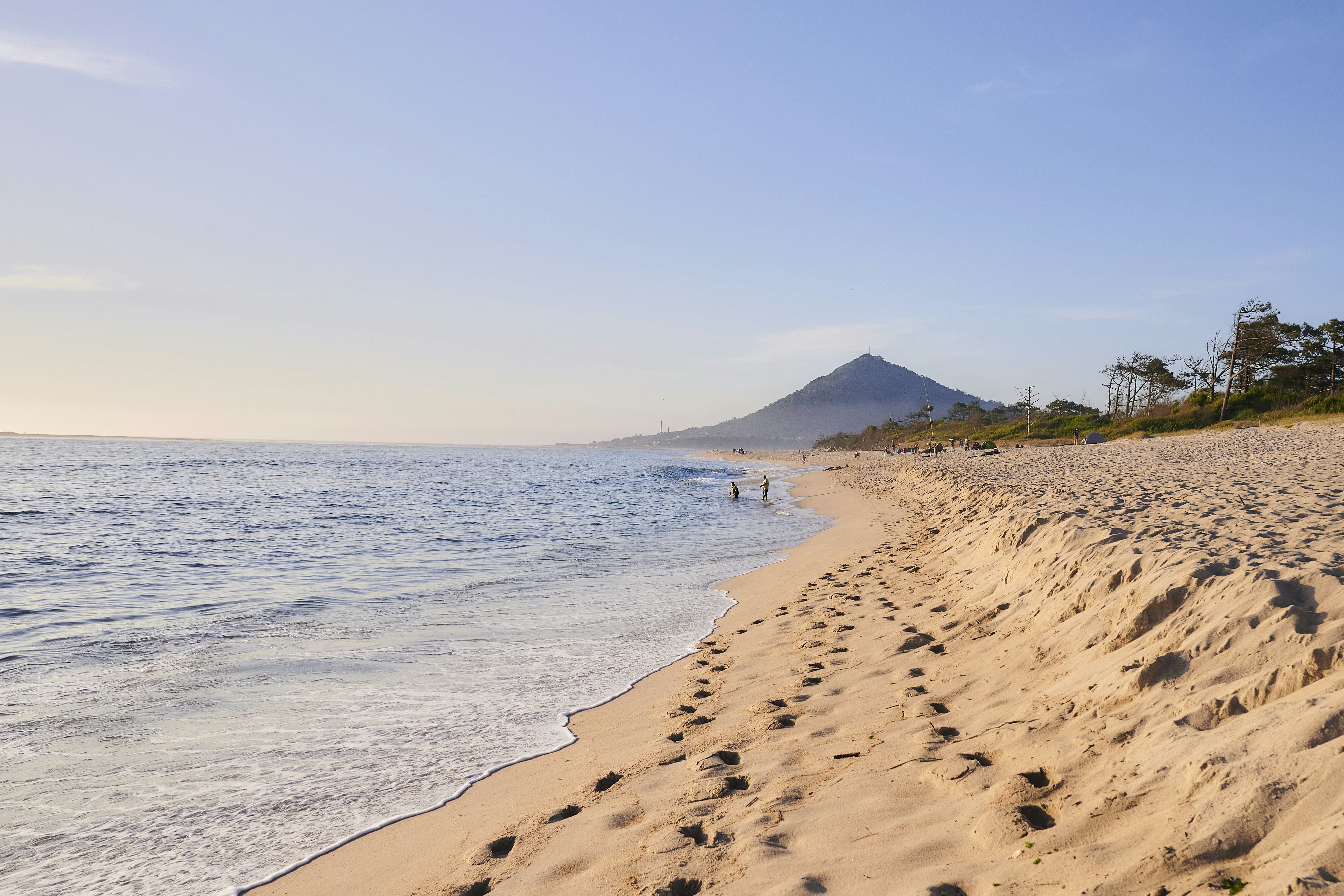 Two people at the edge of the surf at an early morning on a sandy beach. A pointed hill dominates the skyline in the distance.