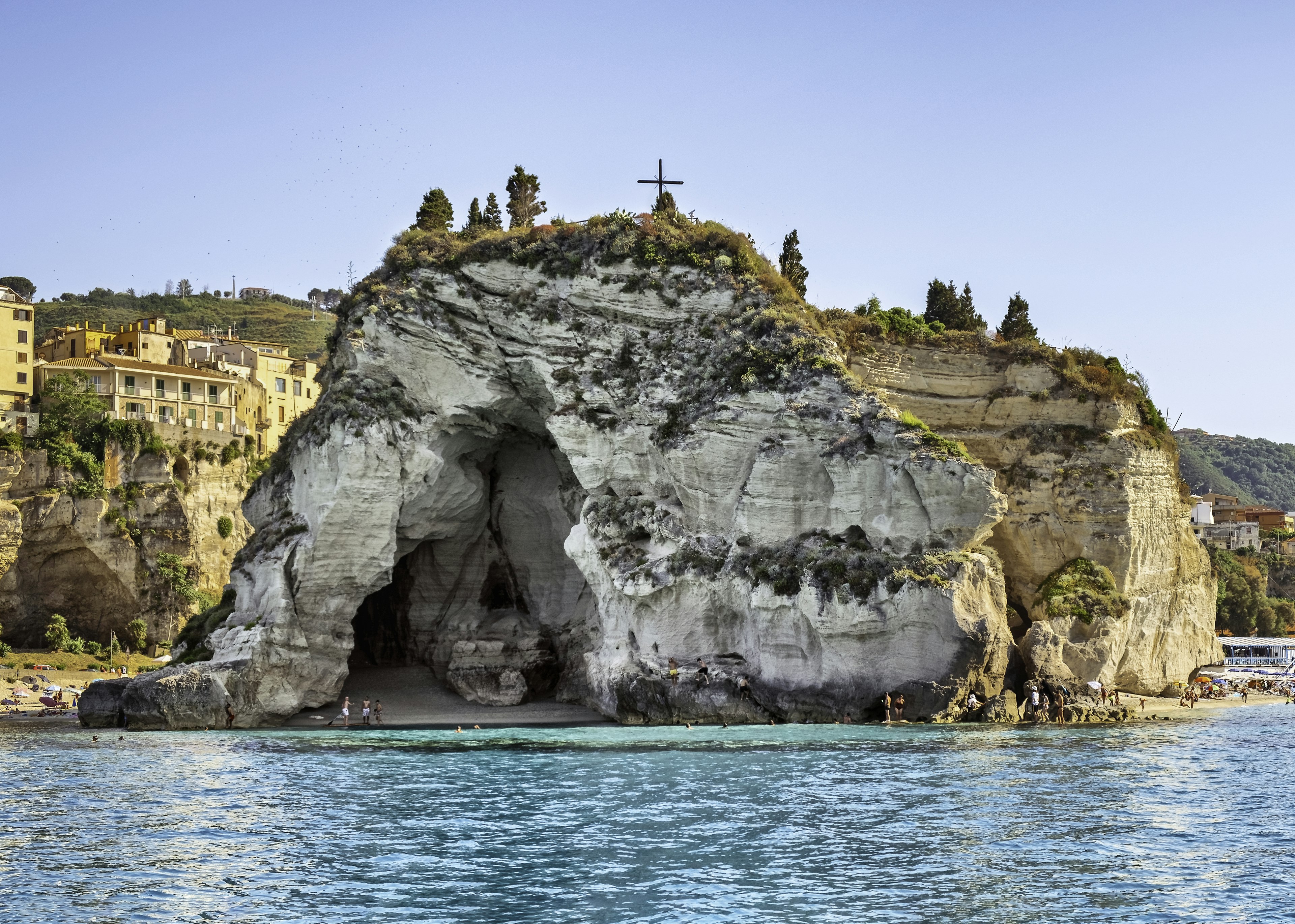 A cave within a cliff with people heading towards the sandy beach.