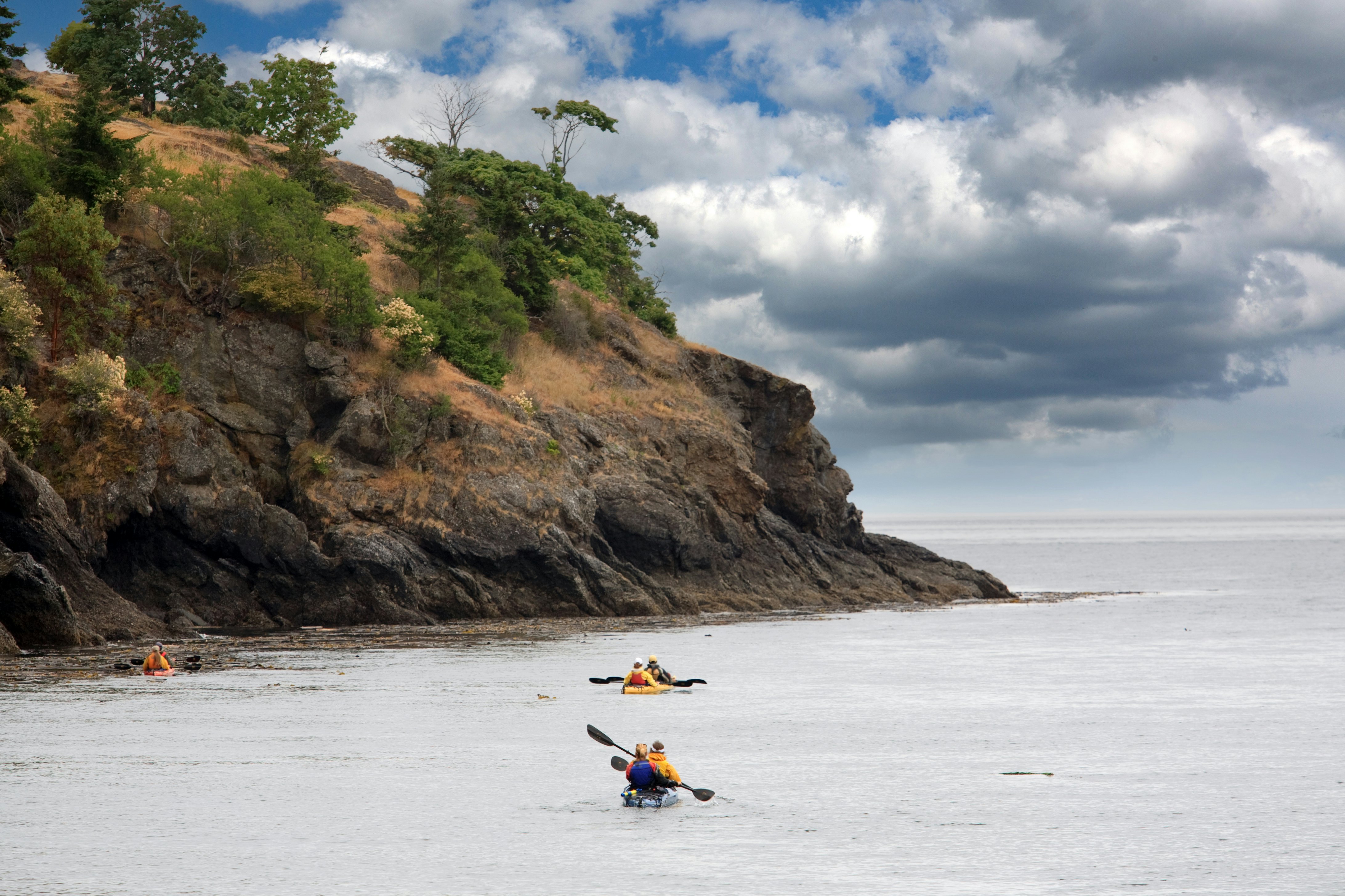 Kayakers in pairs paddle through the ocean near a rocky coastline.