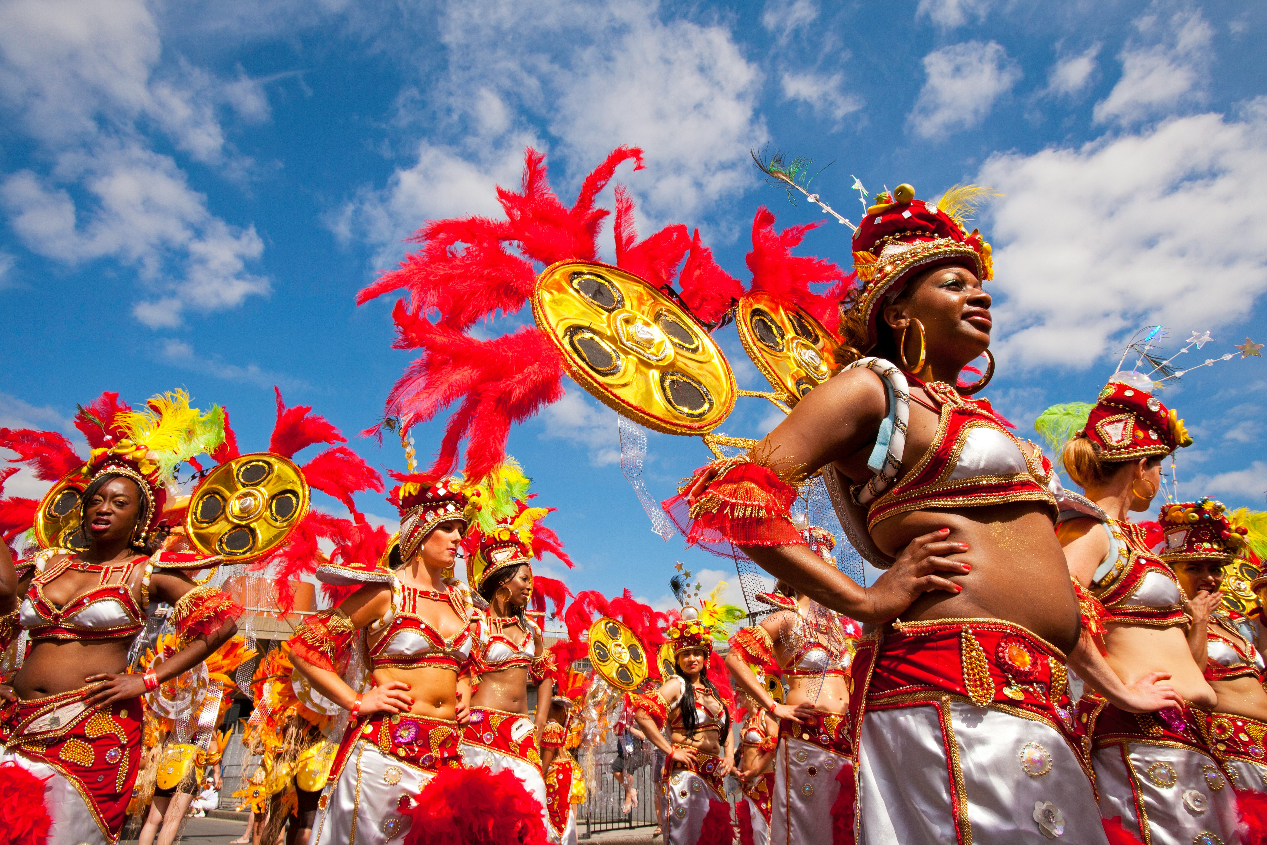A group of women, in matching red and silver costumes with feathered headdresses, get ready to perform at a carnival under blue skies.