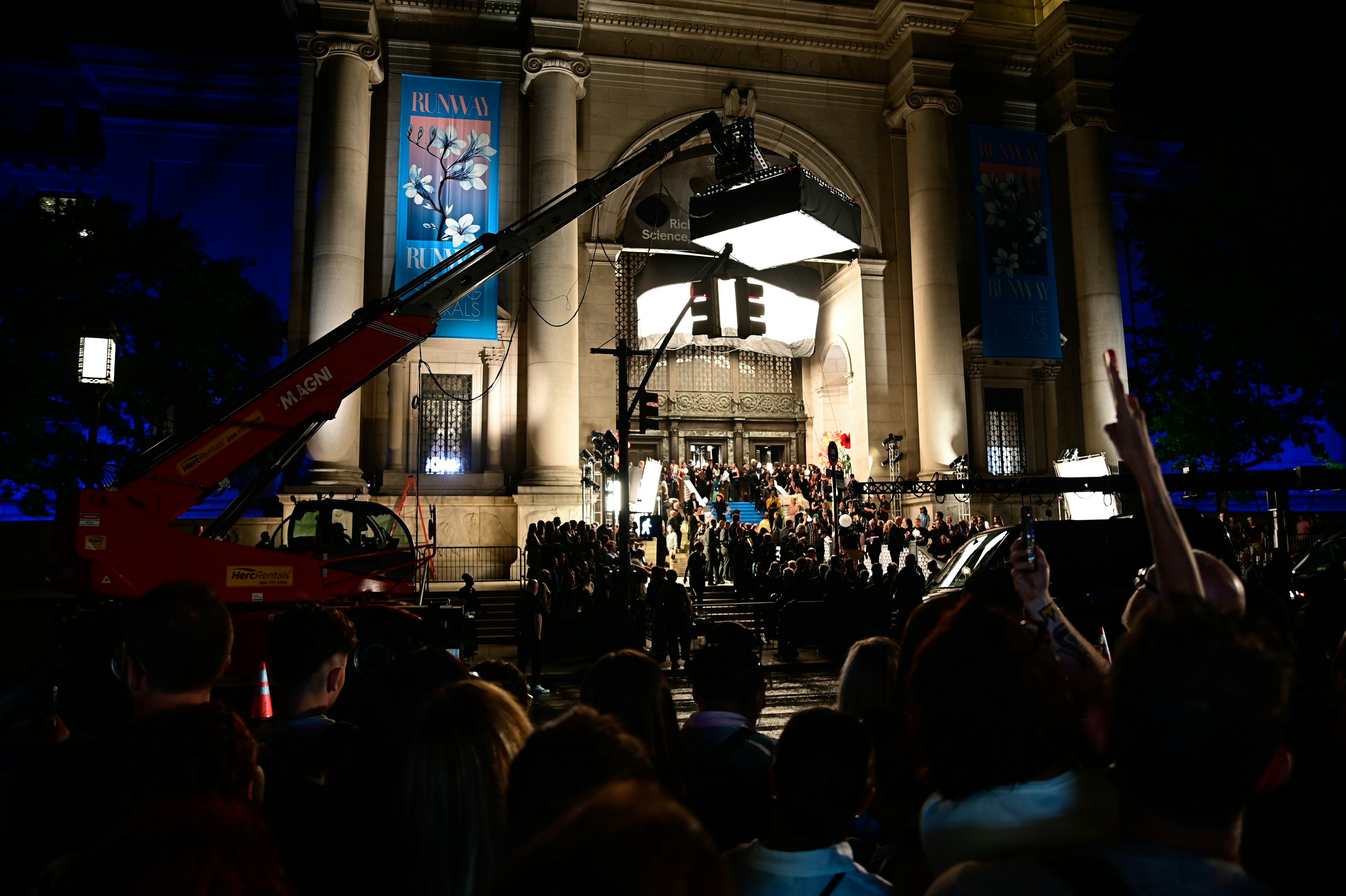 Film equipment is set up in a city street for a nighttime shoot in front of the entrance to a museum.