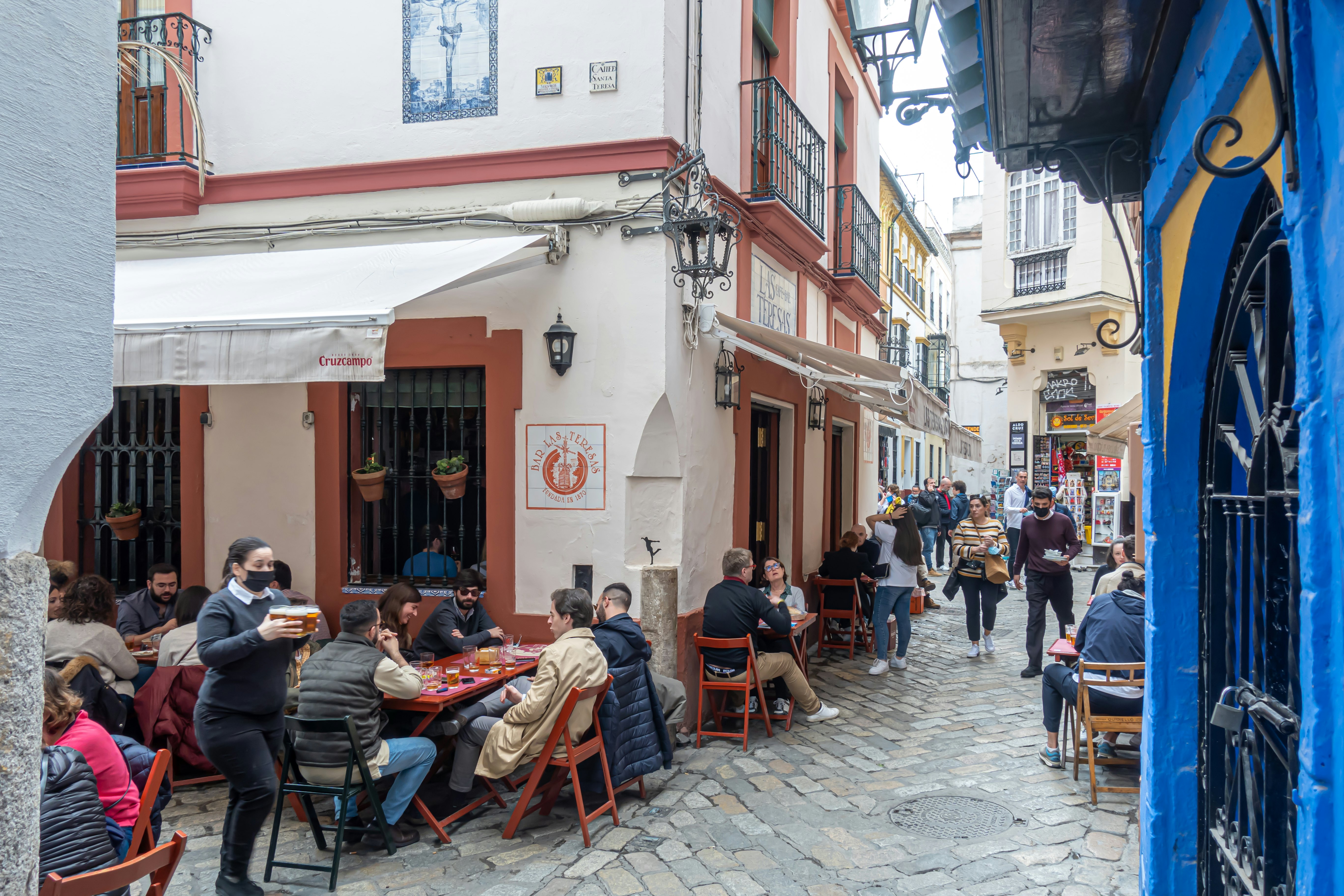 People sit at tables set up in a narrow street outside a restaurant in a historic neighborhood of a city.