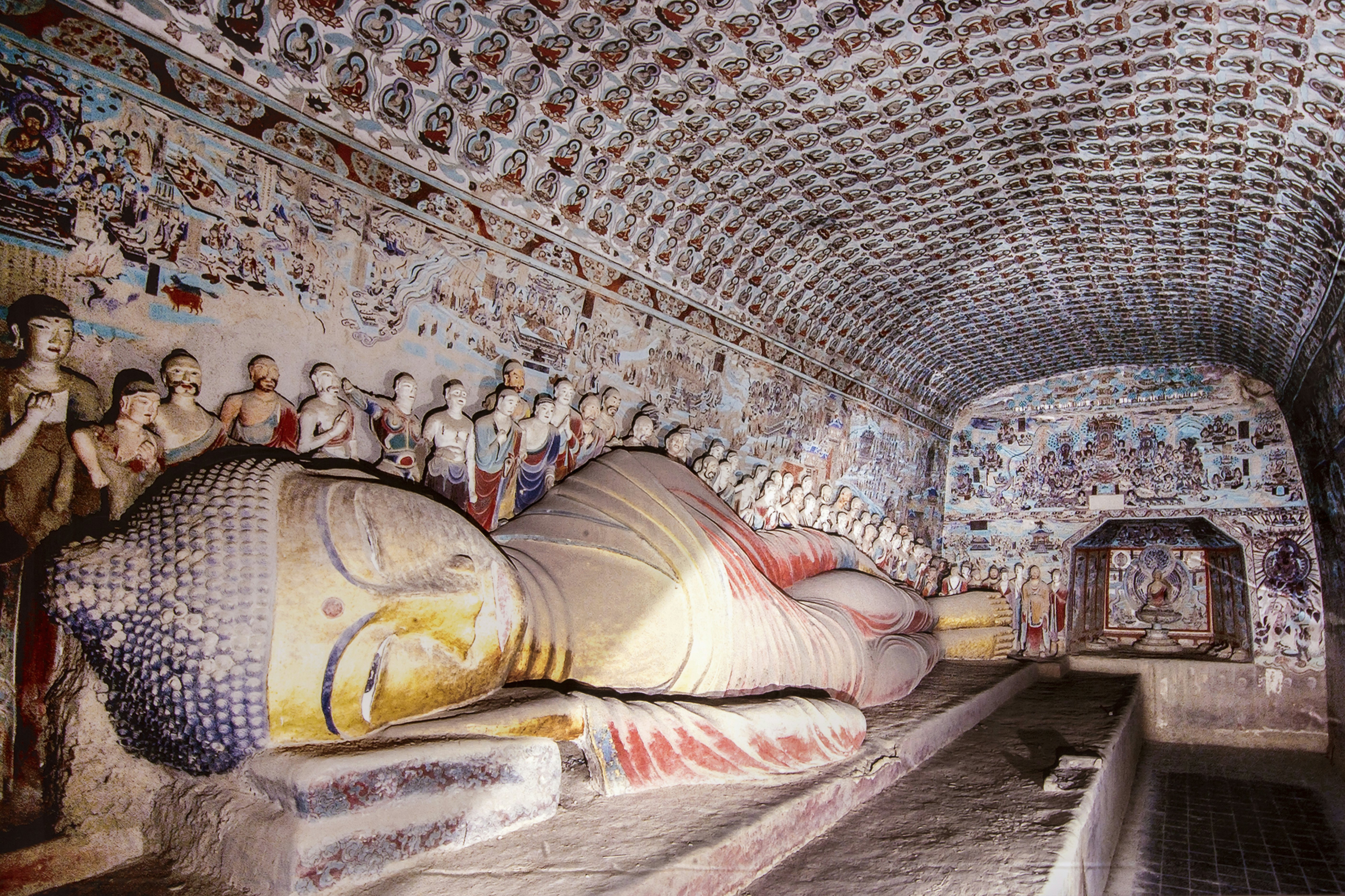 A cave heavily decorated with scultures and wall paintings. A reclining buddha lies across the cave.