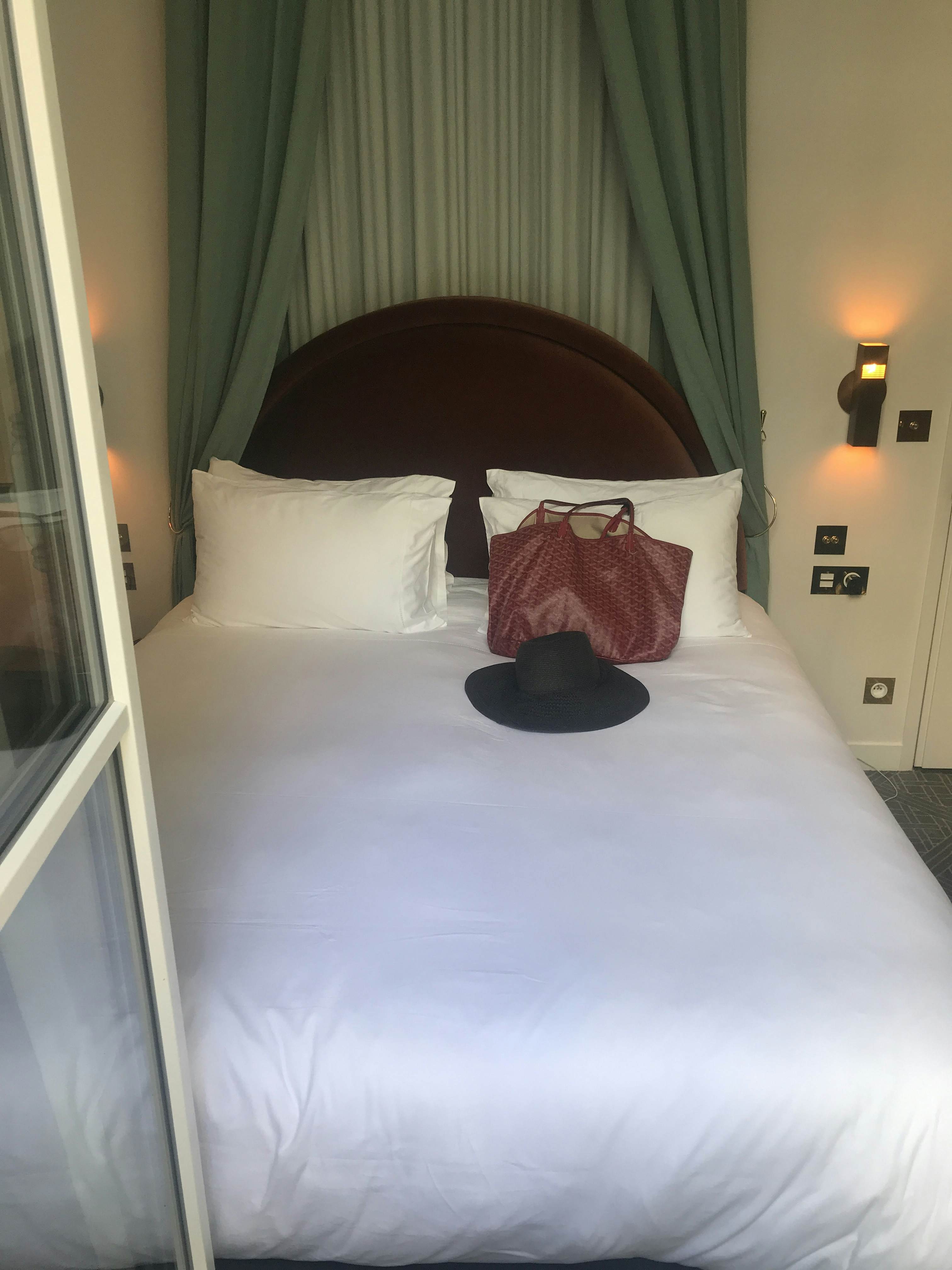 A bed with a white covering and pillows; a black hat and red bag are on the bed.