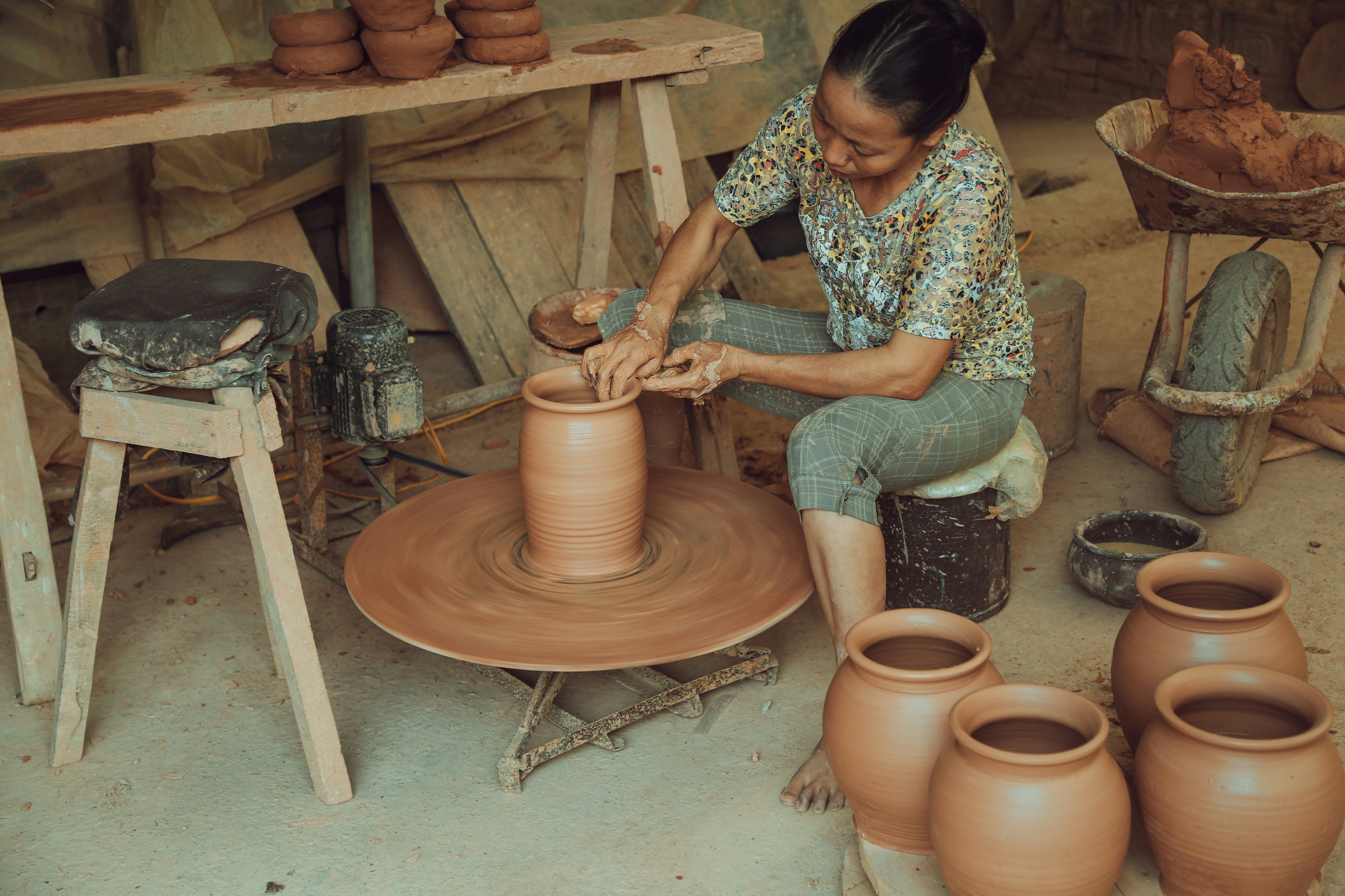 A woman sits and works at a potter's wheel, surrounded by vases