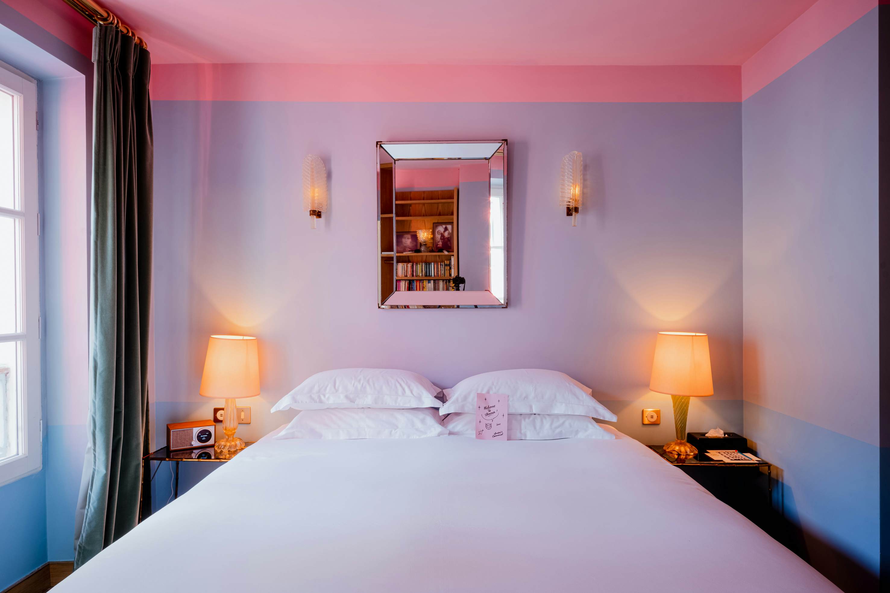A bed with a white covering and four white pillows; lamps are on the bedside tables, and the wall behind the bed is lavender.