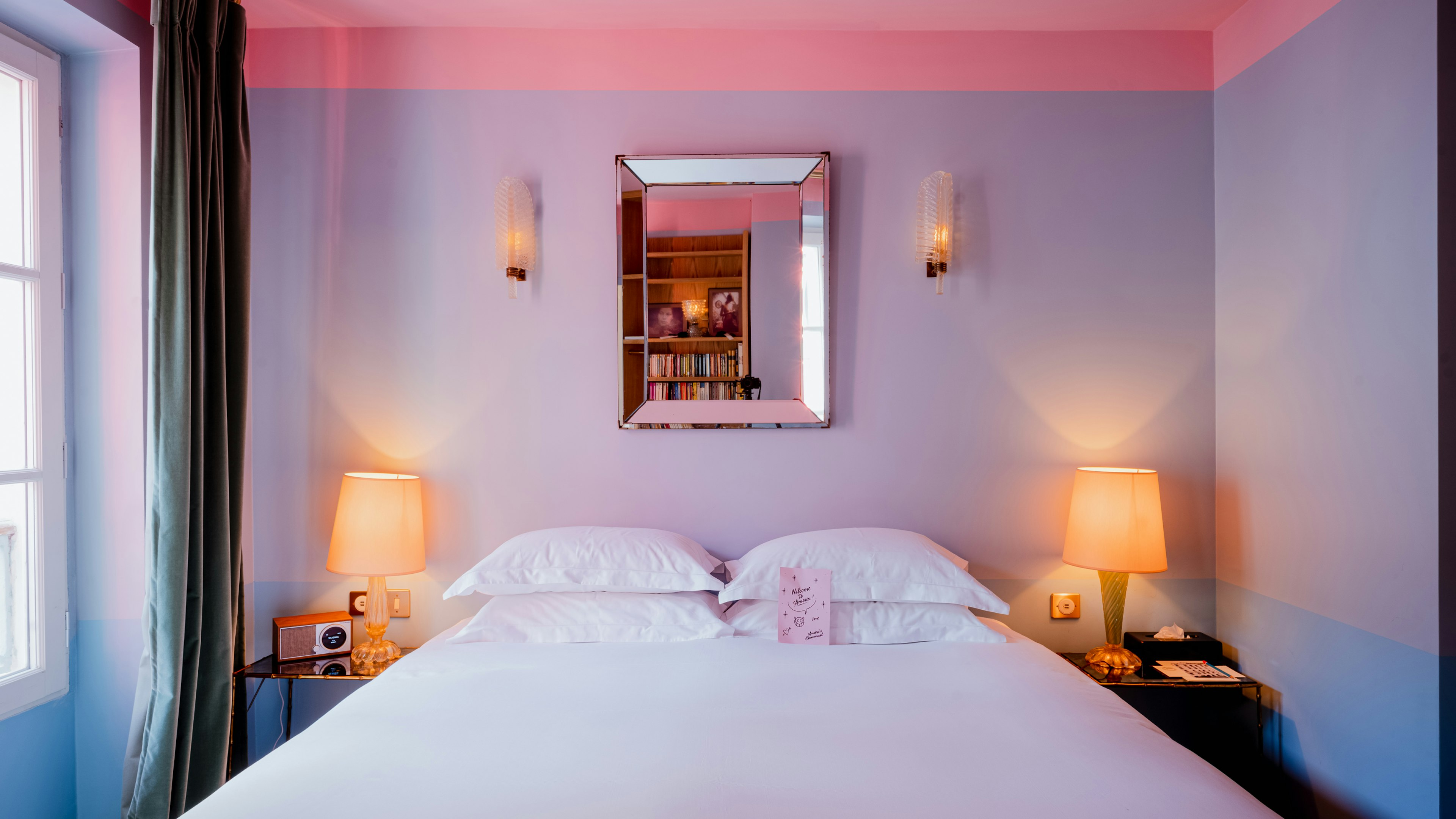 A bed with a white covering and four white pillows; lamps are on the bedside tables, and the wall behind the bed is lavender.