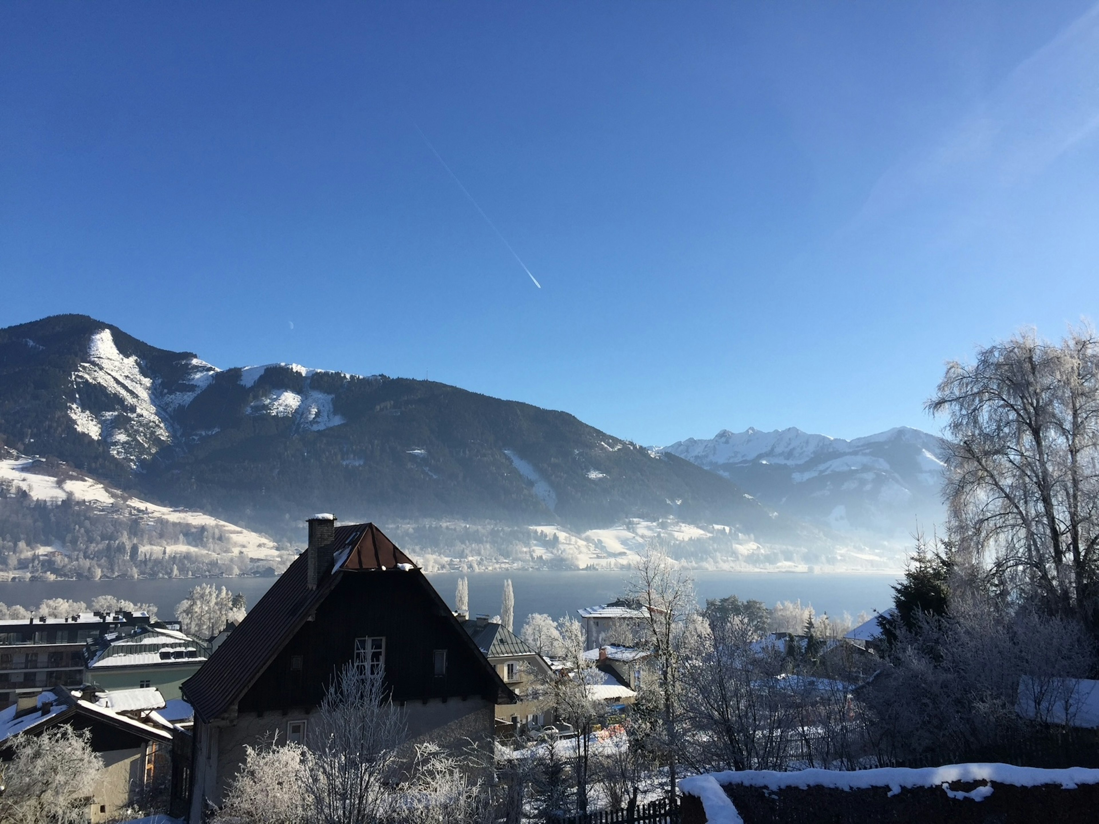 Snow on mountains with evergreen trees; a peaked roof and bare trees are in the foreground.