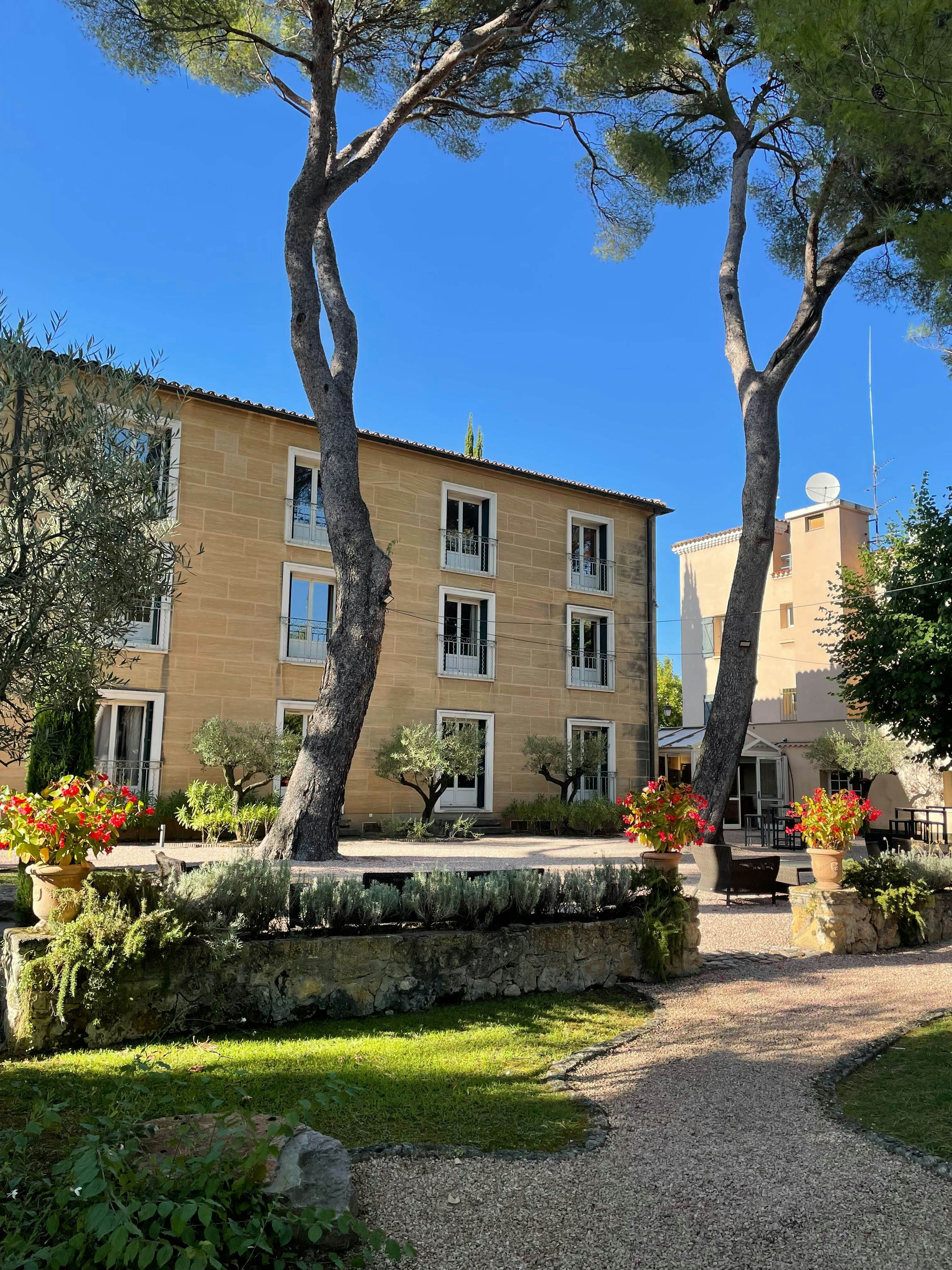 A tan stone building with trees and plants in front of it.