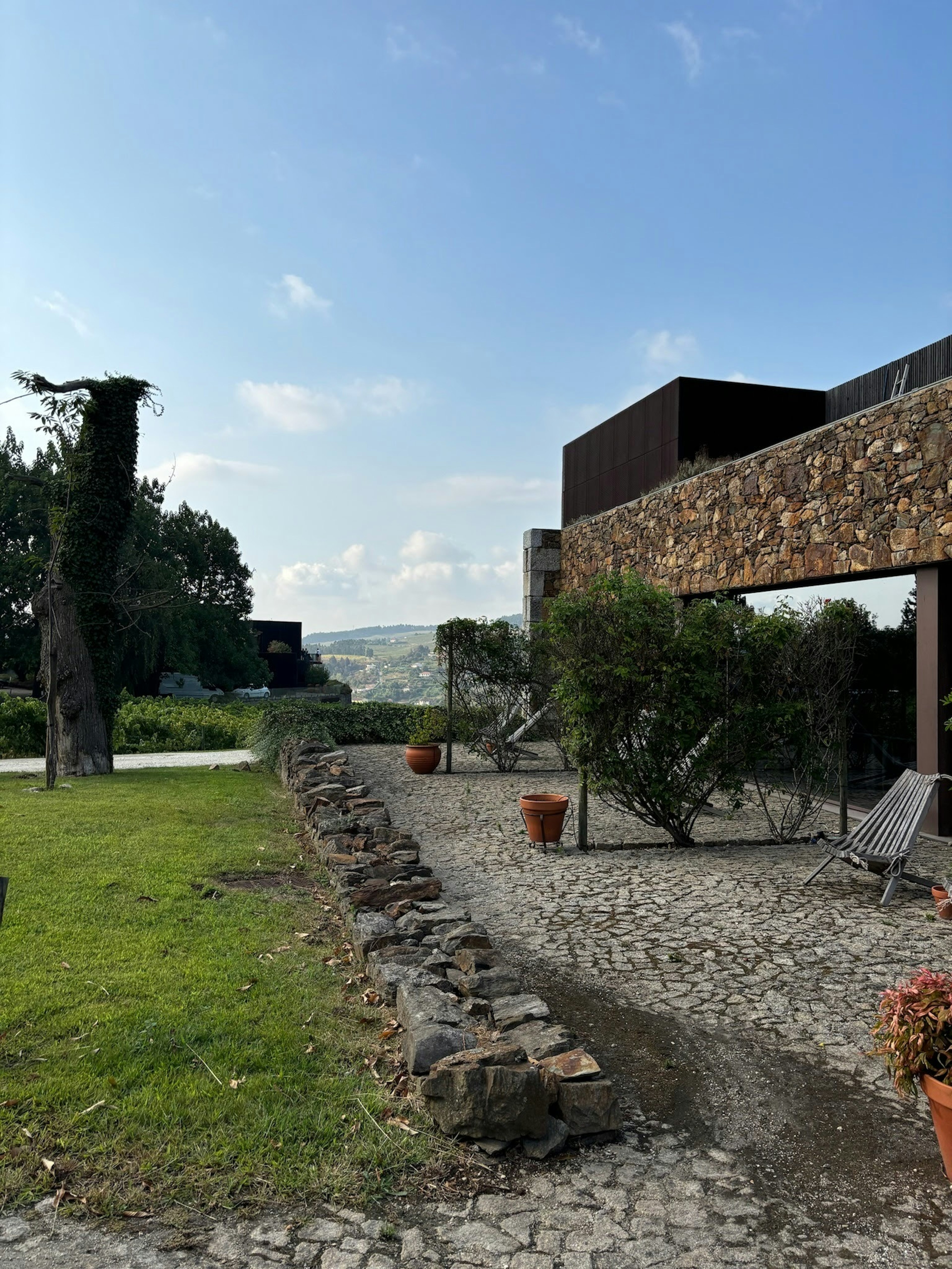 A cobblestone patio has wooden chairs, a few terra-cotta planters, and a bush; on the other side of a low wall is green grass.