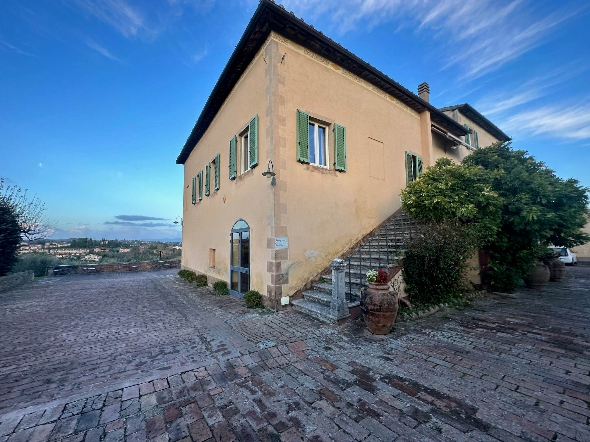 A staircase leads up the side of a tan building with green shutters. A cobblestone patio surrounds the building, and there are distant views of the countryside.