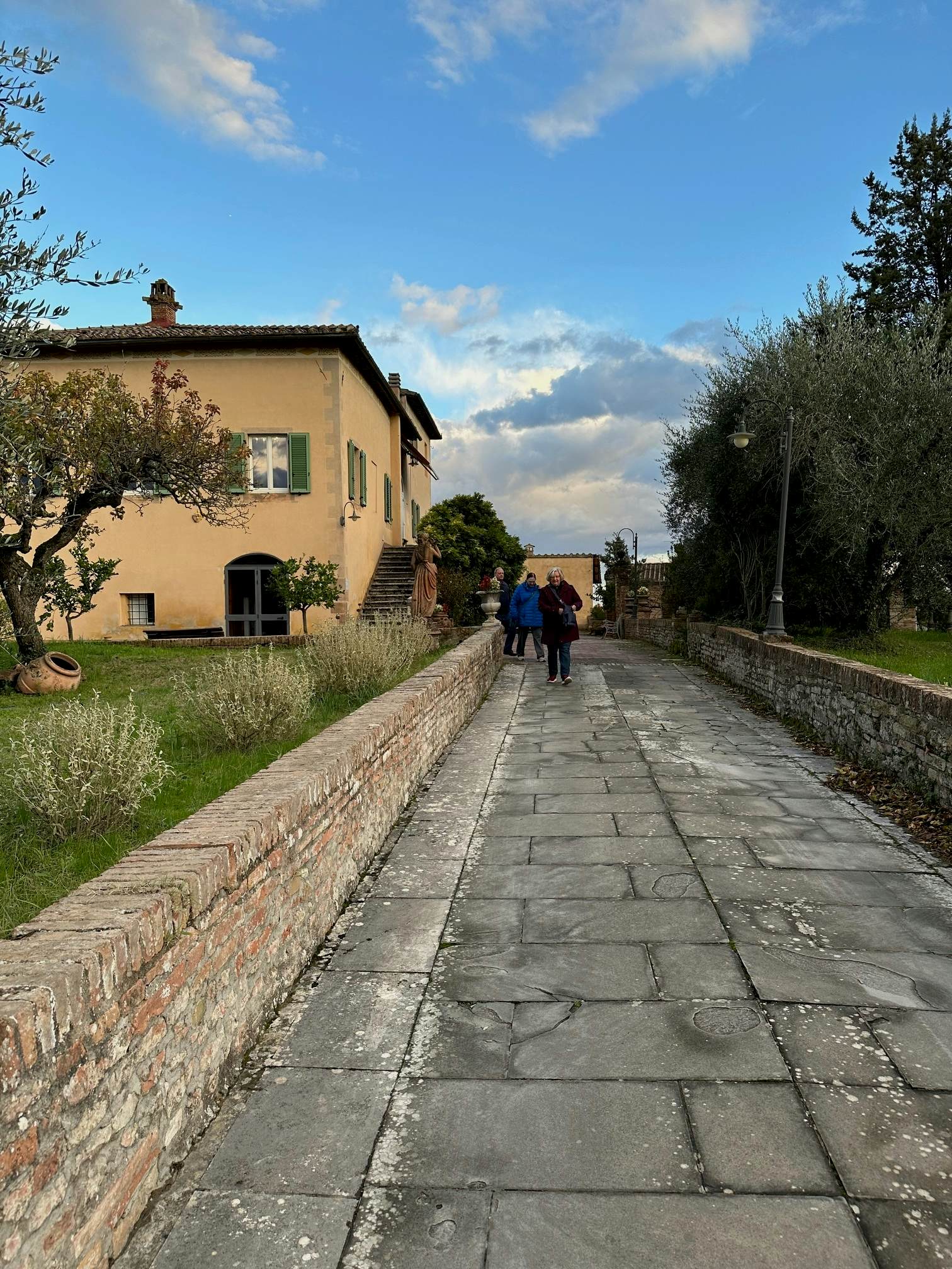 A cobblestone walkway has a low stone wall to one side; a tan building with green shutters is ahead on the left side of the wall.