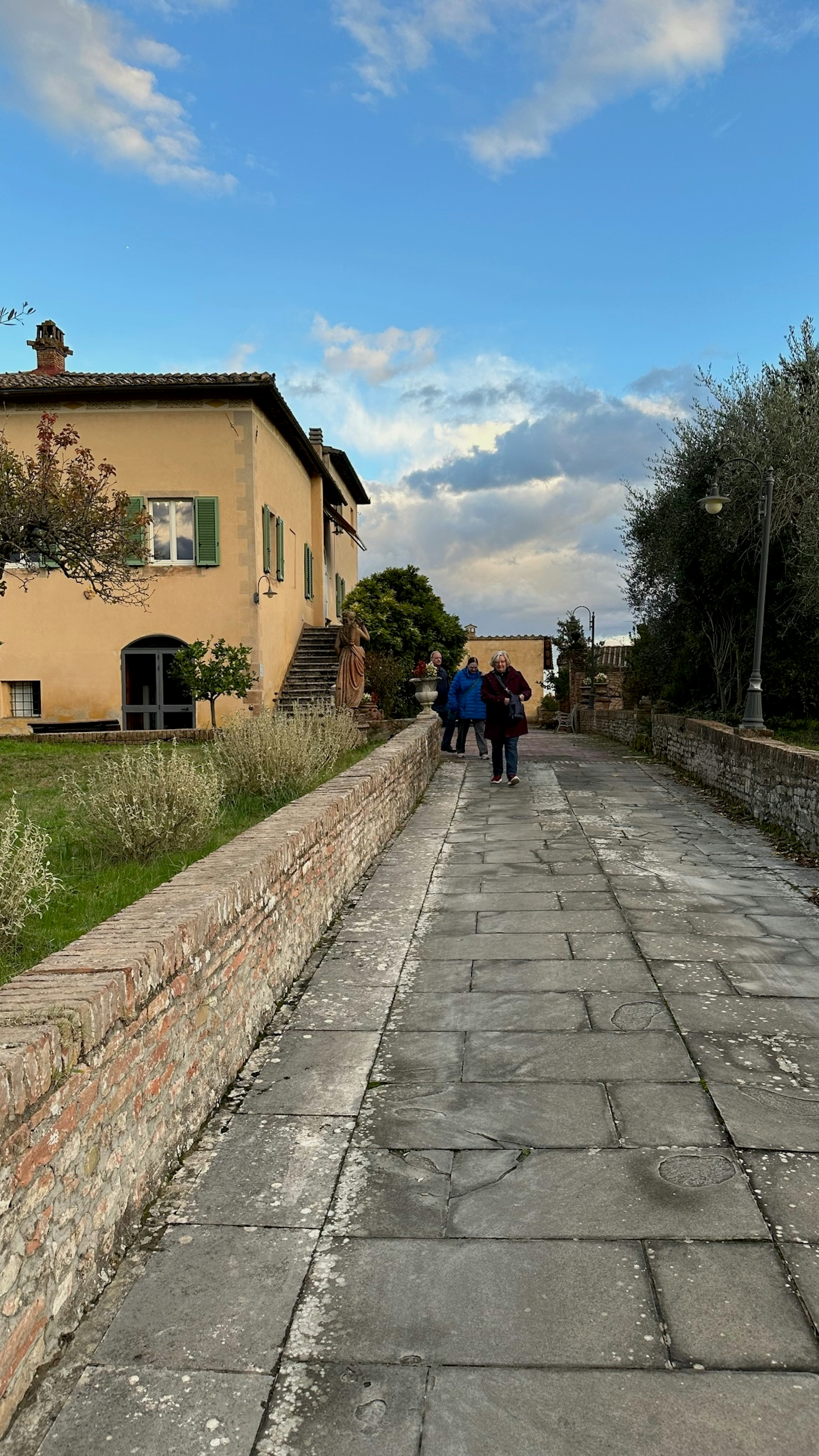 A cobblestone walkway has a low stone wall to one side; a tan building with green shutters is ahead on the left side of the wall.