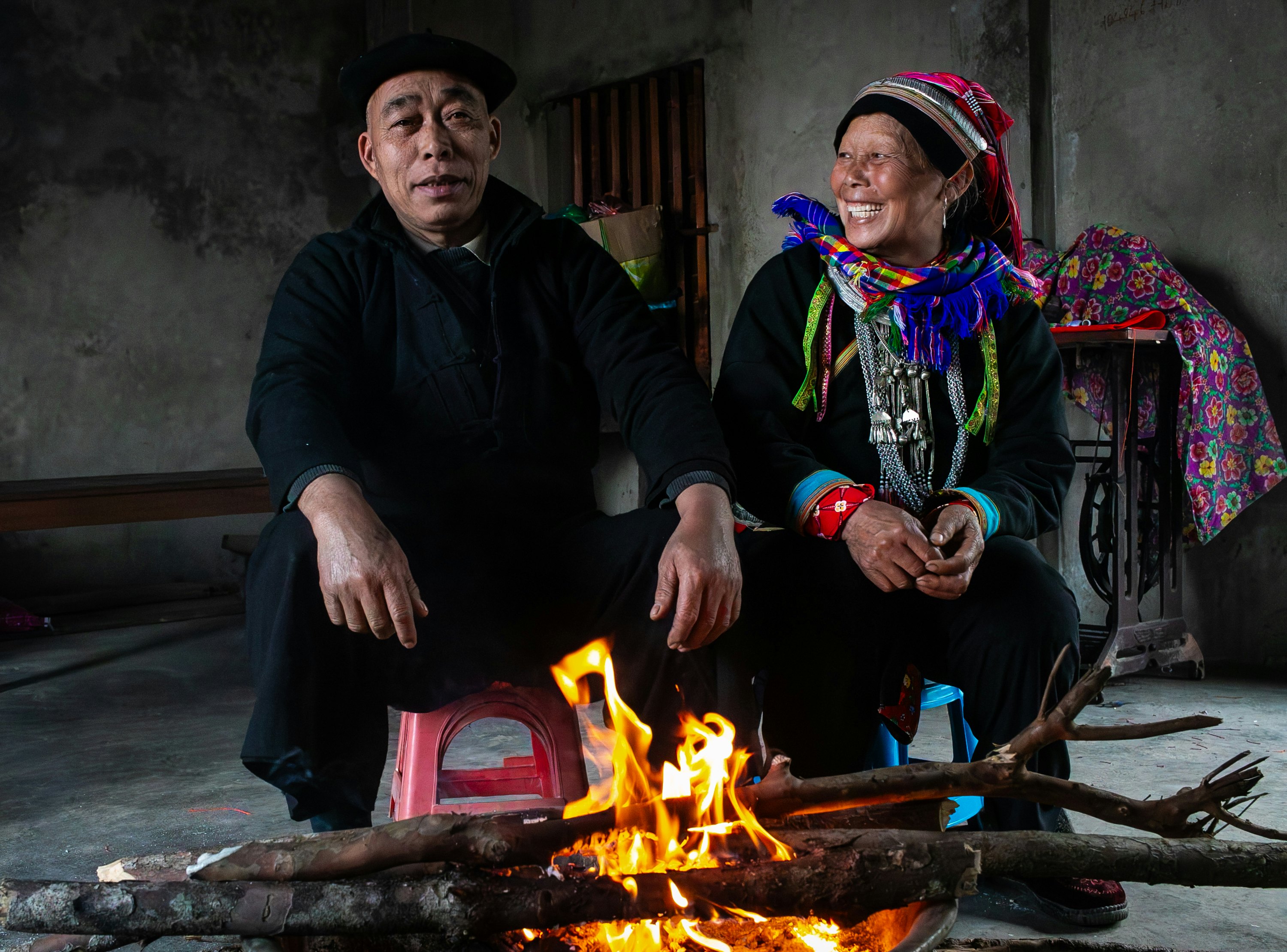 A man and woman sit on stools in front of a small fire. She is wearing colorful woven accessories.