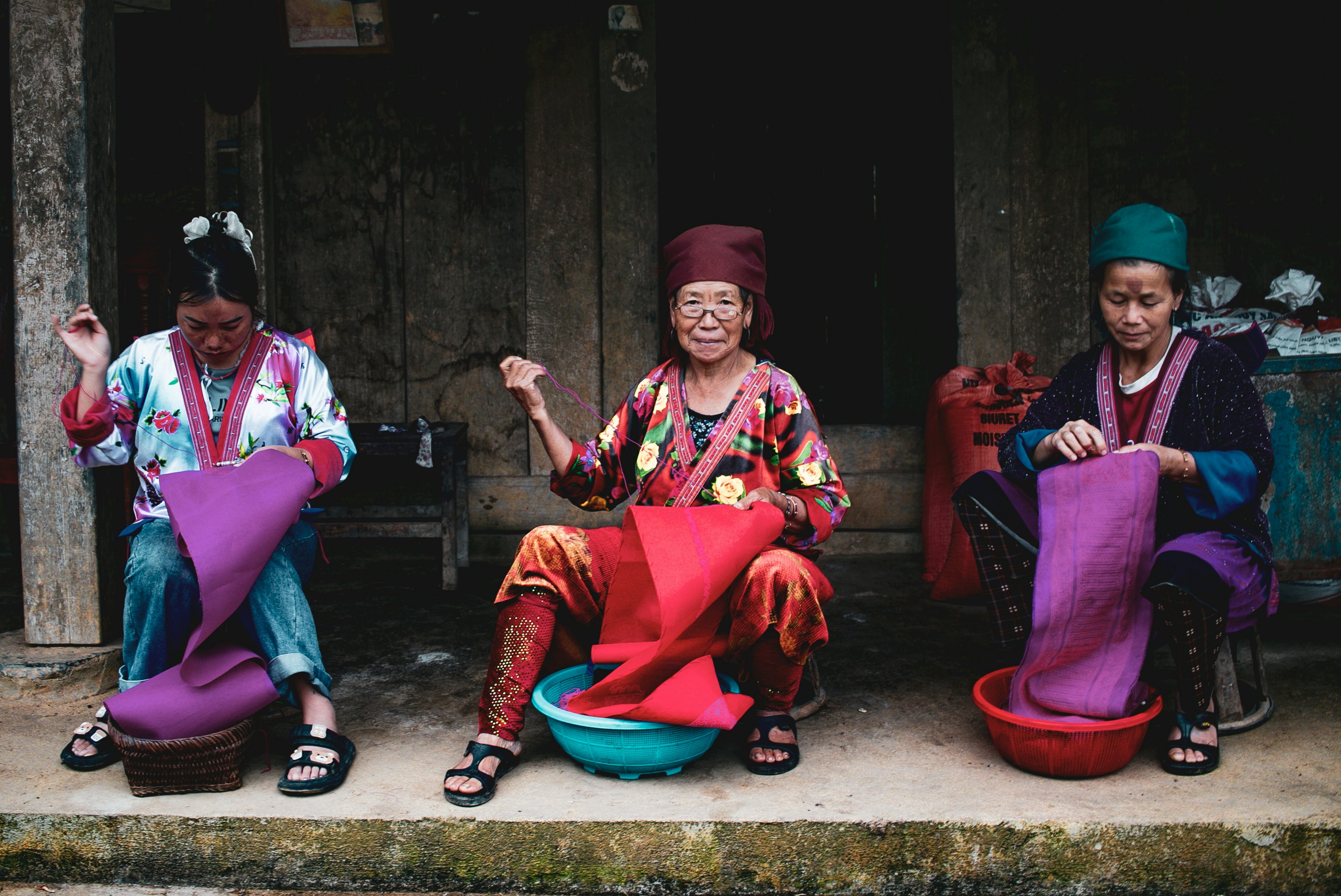 Three people are seated with baskets between their feet while sewing colorful fabric.