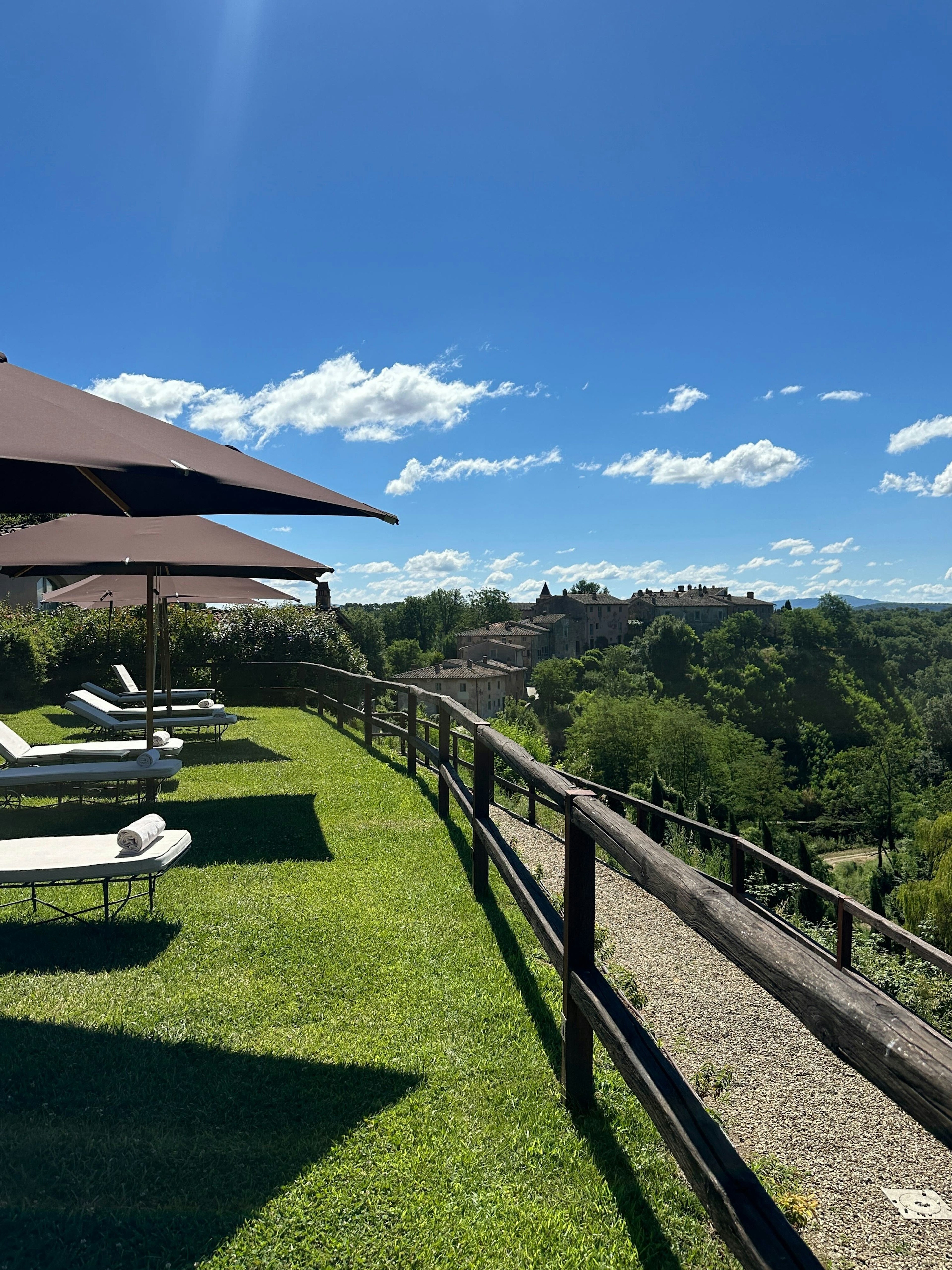 Lounger chairs in grass with umbrellas face a forested area; stone buildings are in the distance.