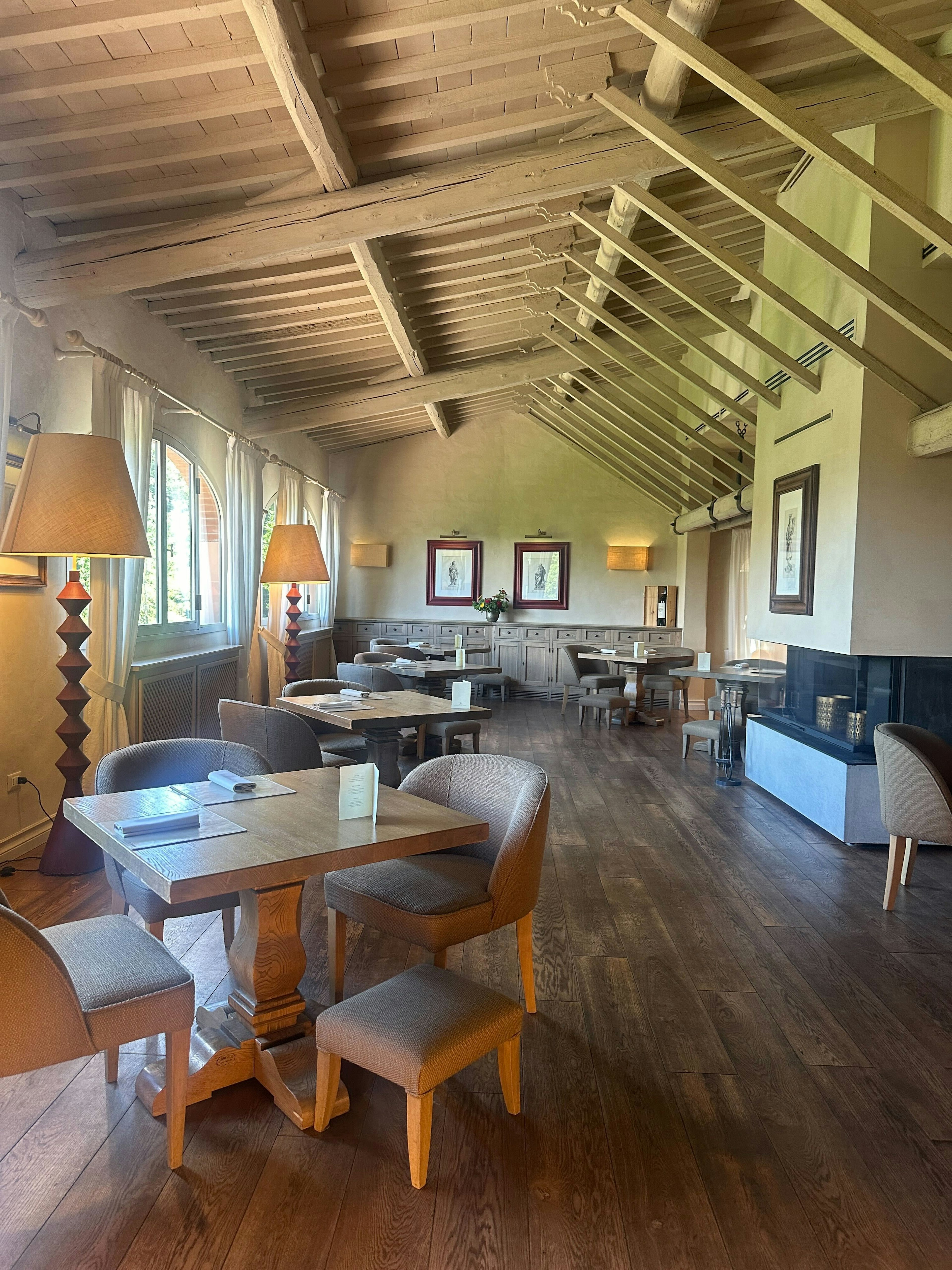 Empty square wood tables, each set with a card, folded napkin and two or three chairs and a stool, in a long room.