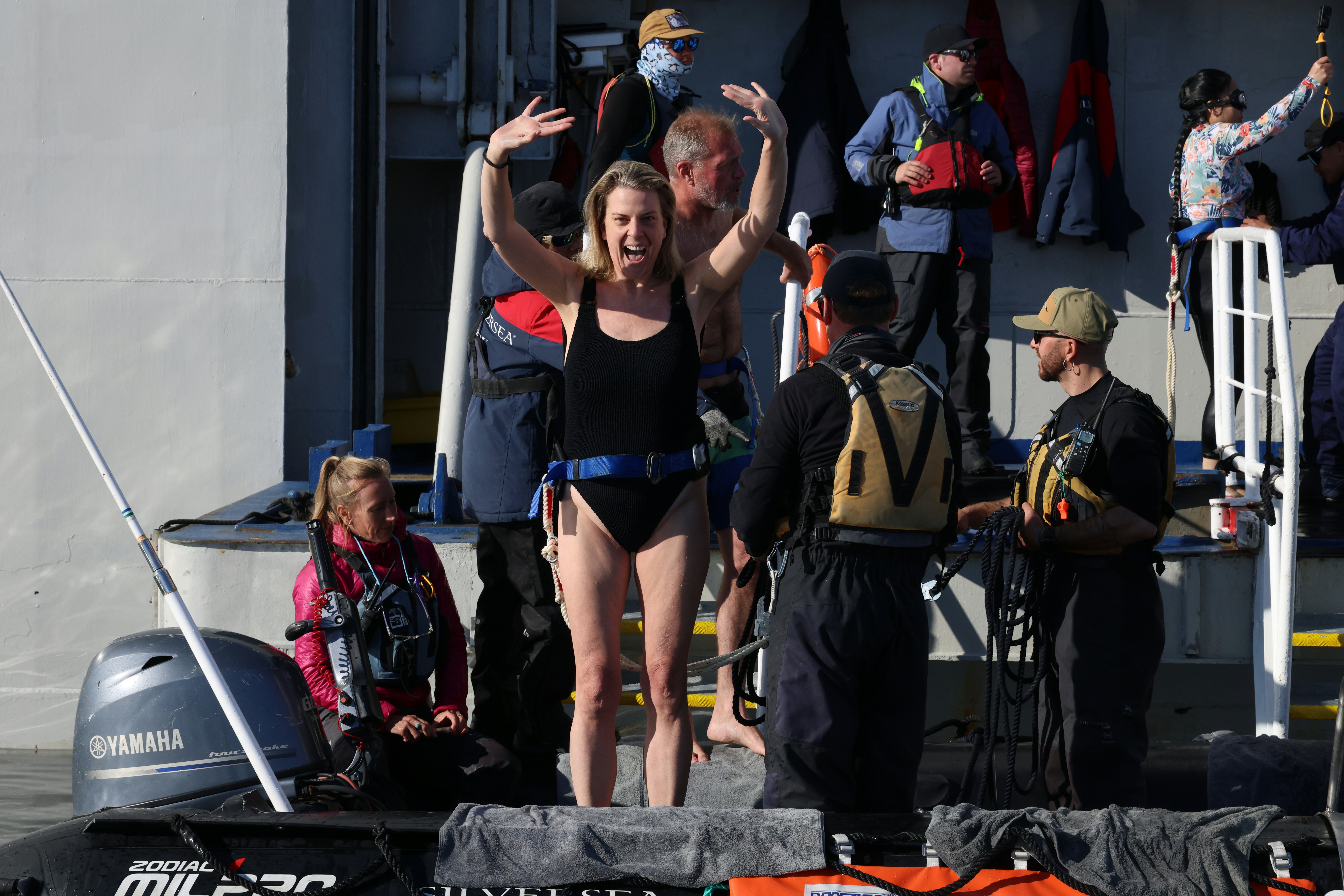 A woman in a swim suit attached to a boat by a waist belt raises her hands and smiles at the camera as she prepares t o jump into the ocean.