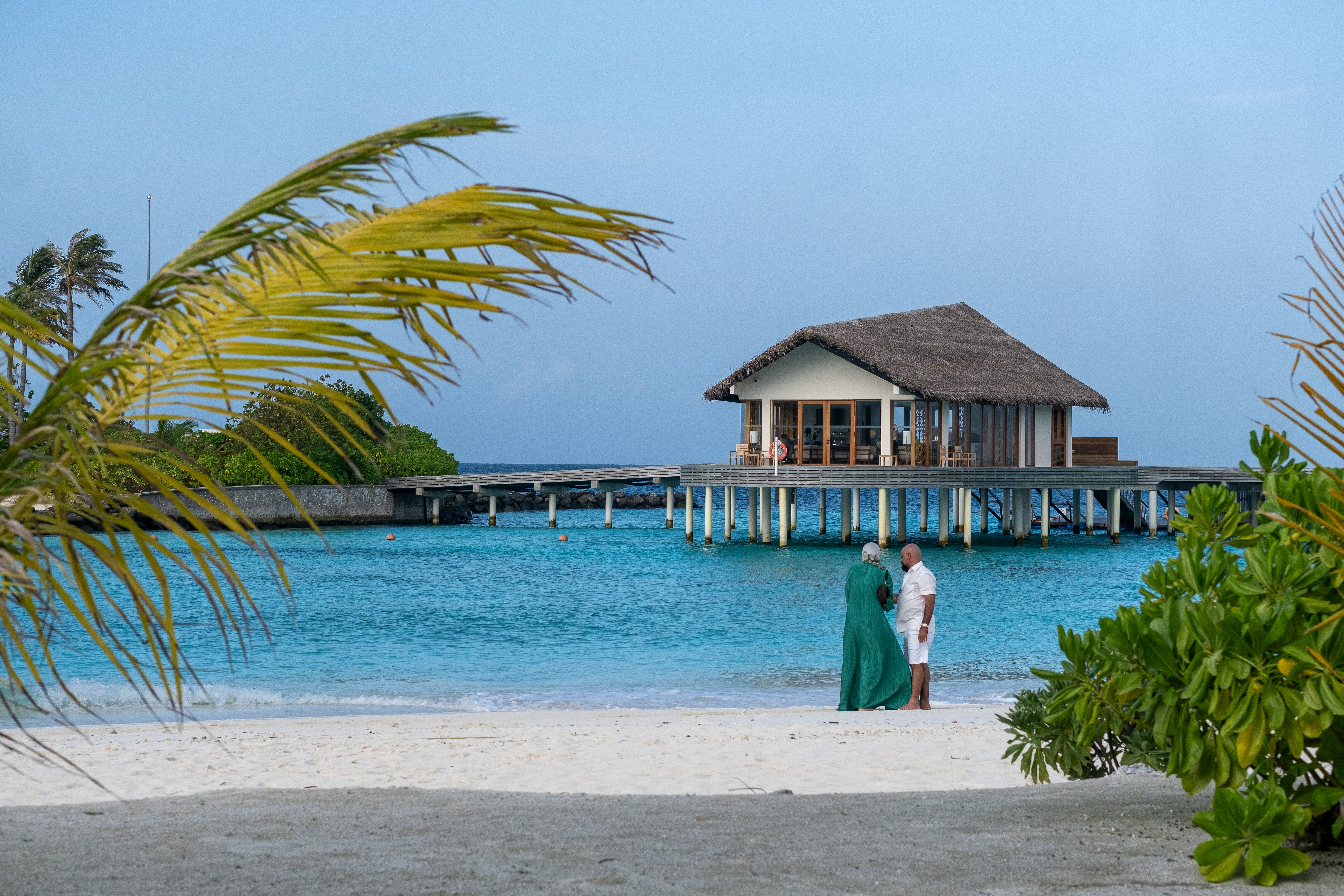 A couple stands on a white sand beach by blue water; a villa is built on stilts over the water. Palm fronds and green bushes are on either side of the sand.