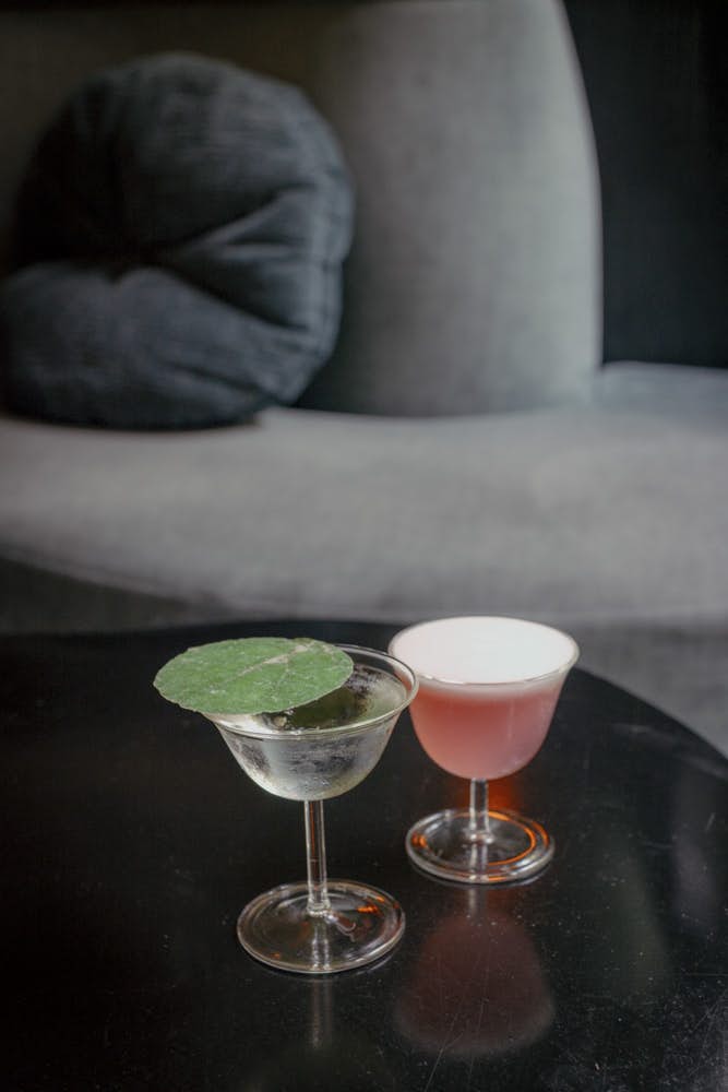 Two clear glasses on a black table; one has a green leaf on top and the other is filled with pink liquid.