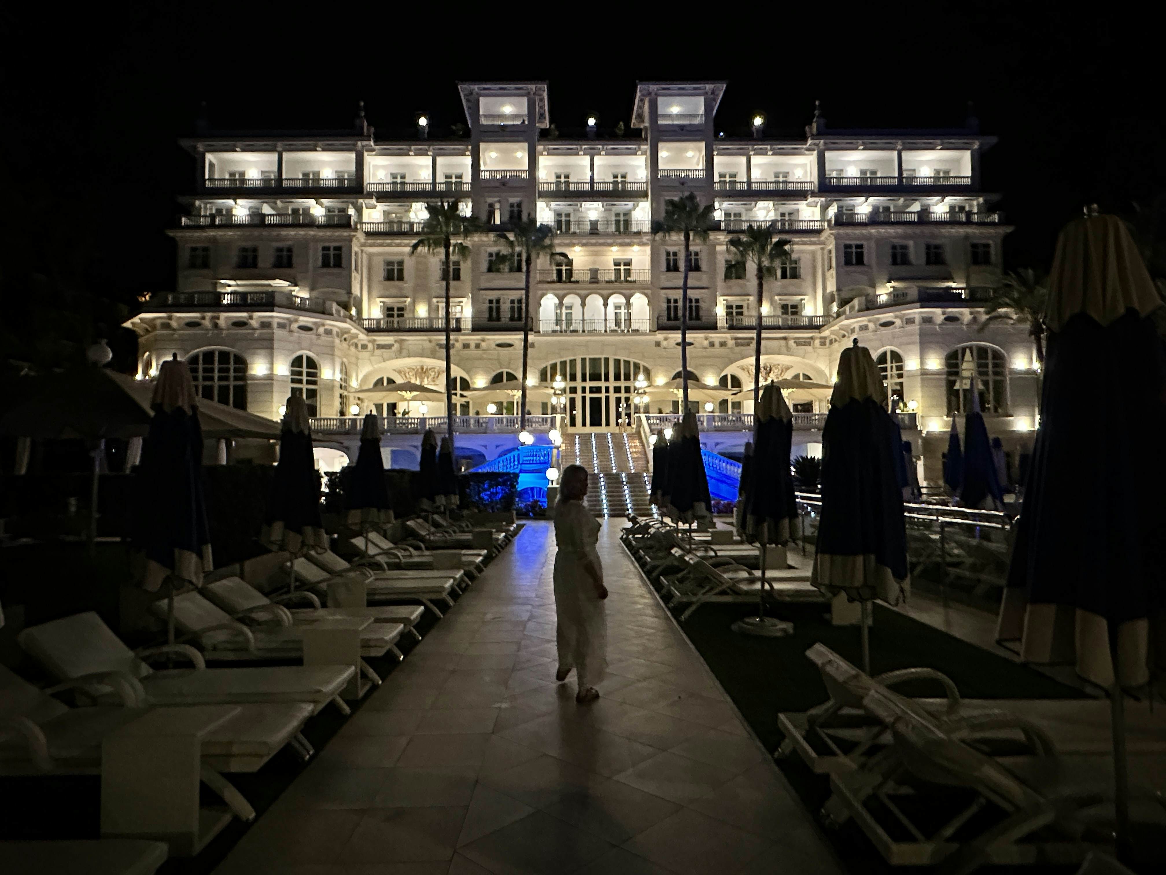 A person on a walkway between empty lounge chairs; a white building lit up at night is in the background.