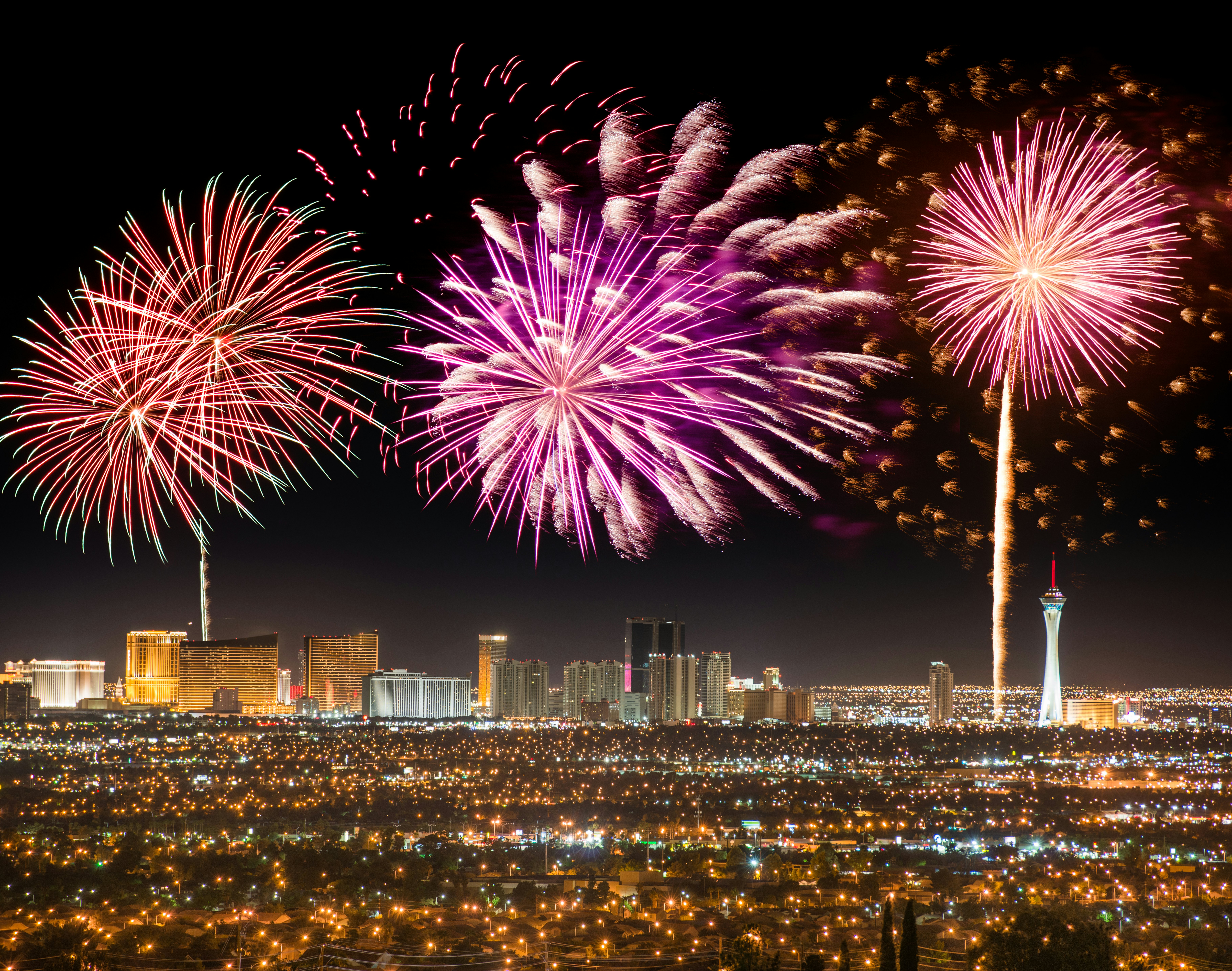A wide shot of huge fireworks exploding over a series of tall hotels and towers at night in a city.
