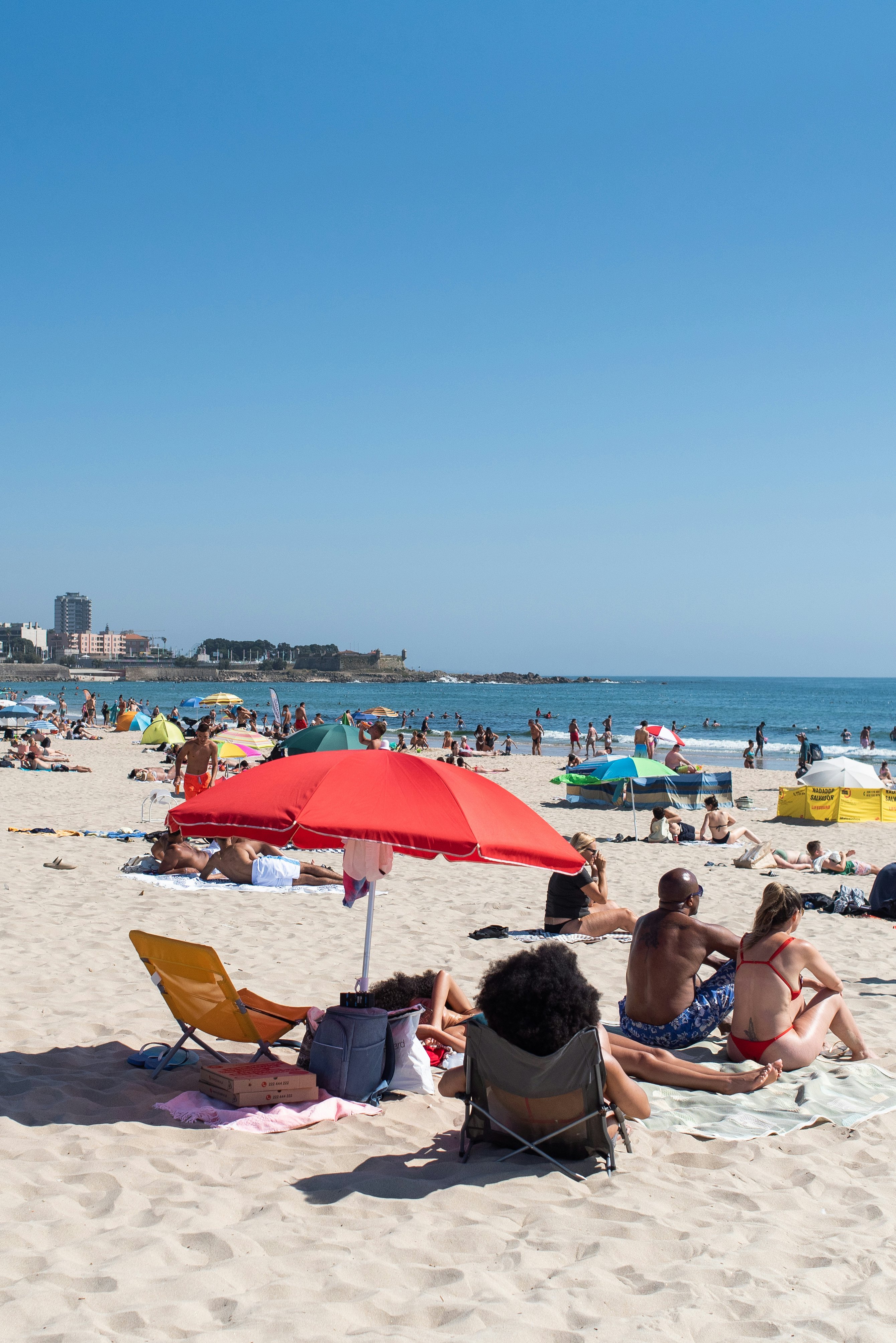 People relax under a red umbrella that is standing on the sand on a busy city beach on a sunny day.