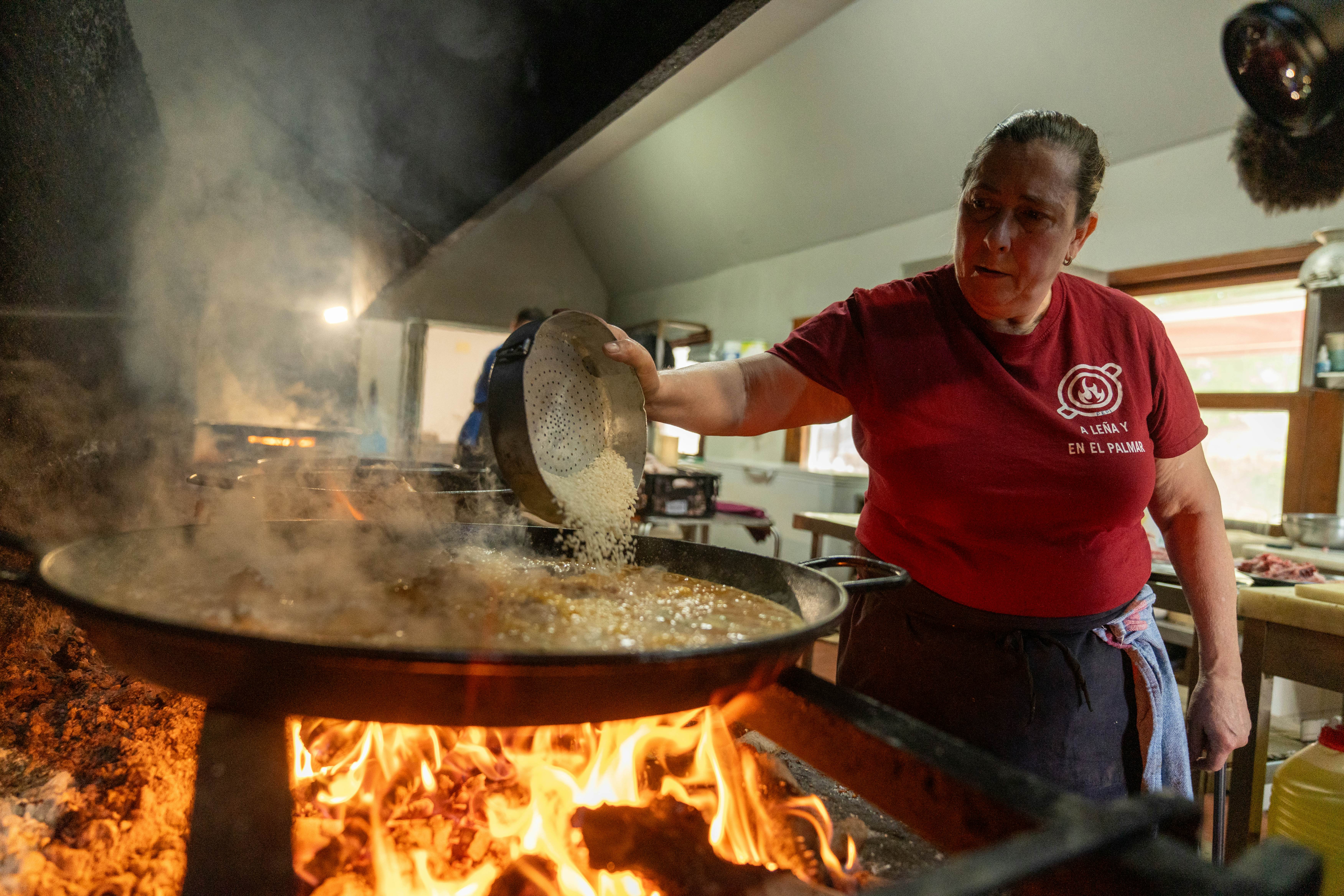 A chef wearing a red t-shirt pours rice into a large flat pan over fire.