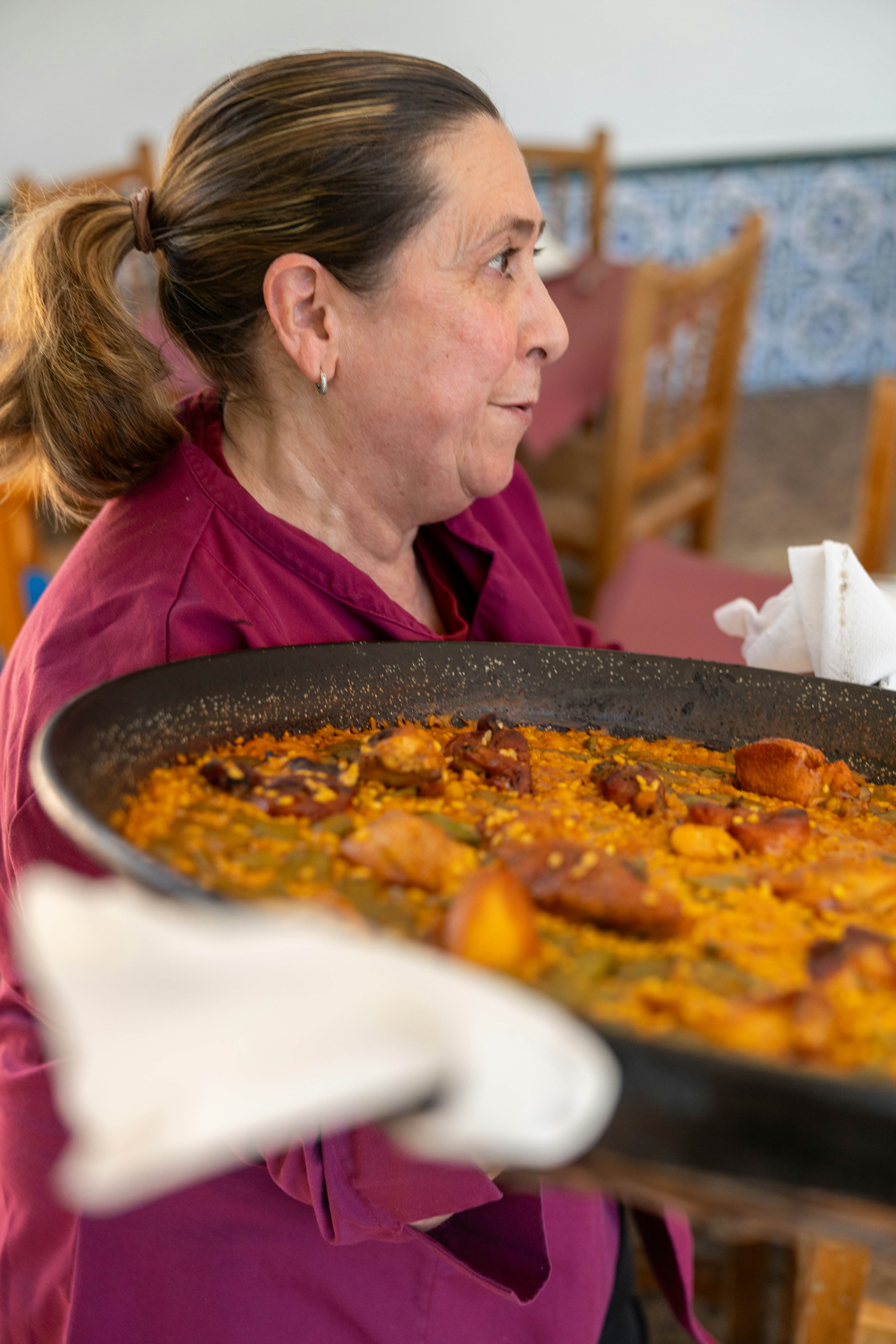 A chef wearing a red t-shirt carries a large pan of paella over her shoulder.
