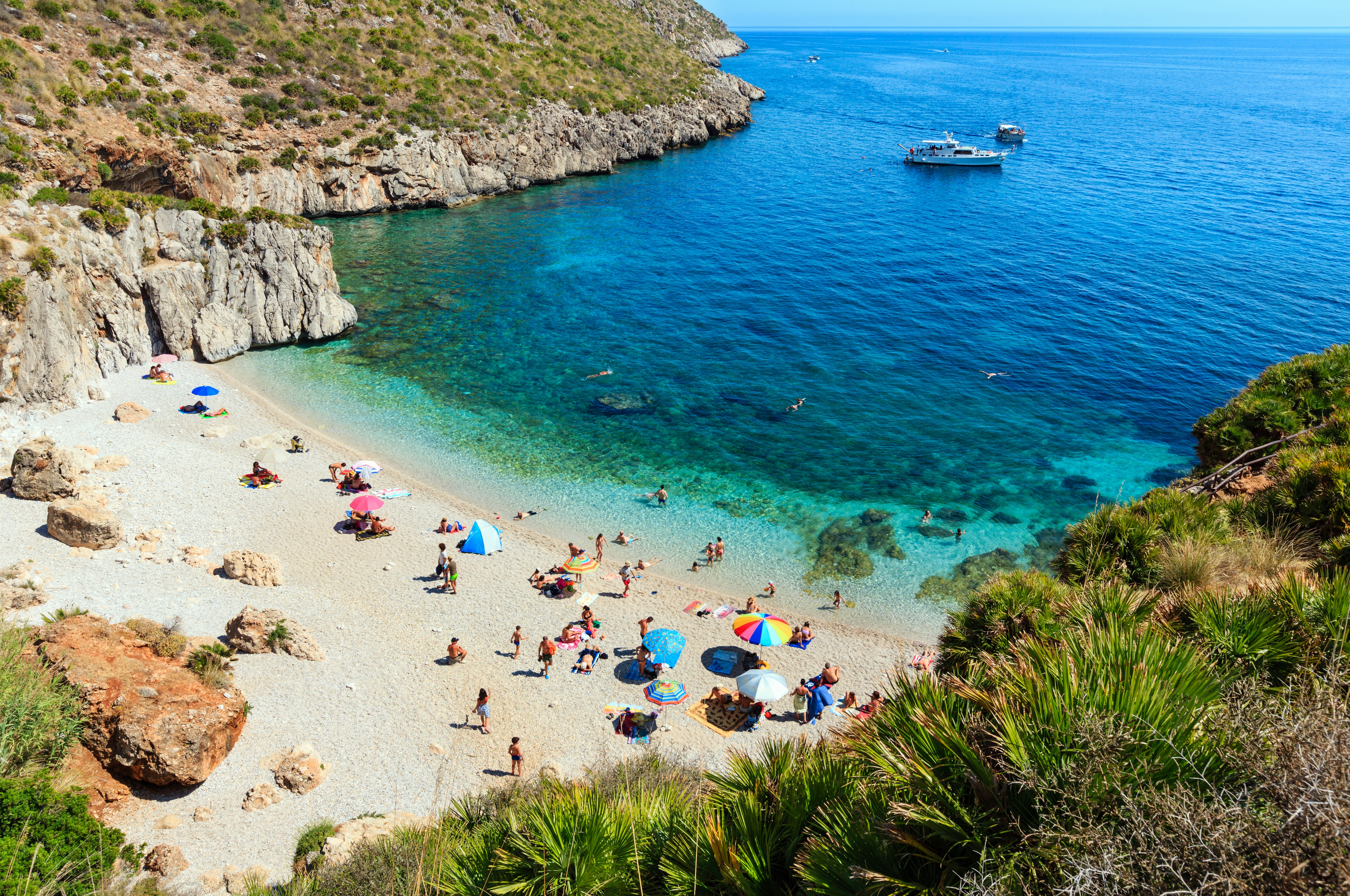 An uncrowded beach in Sicily, Italy, with a few families under beach umbrellas, and a boat in a blue cove.