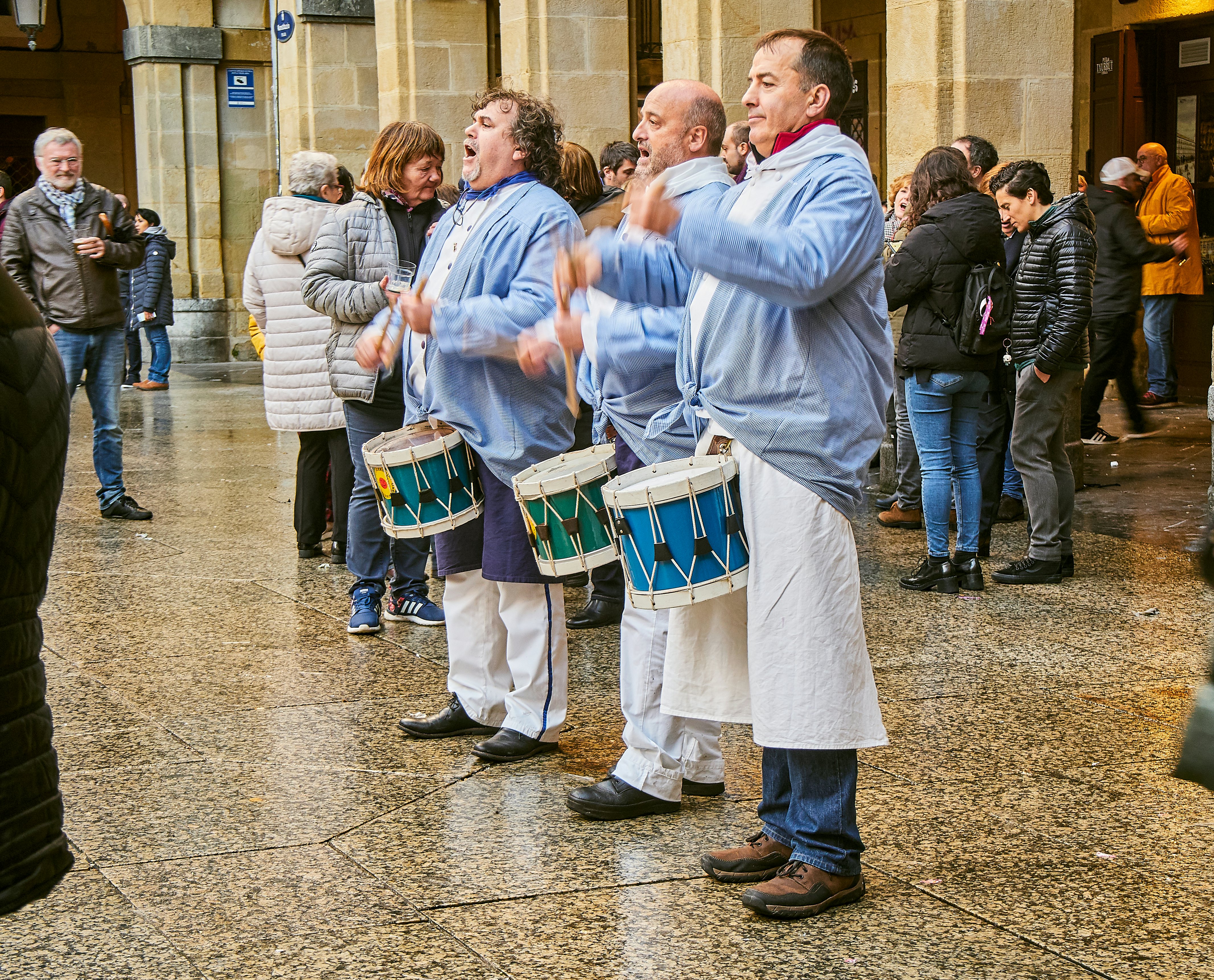 Three drummers wearing blue shirts perform outdoors with people milling about.