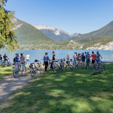 ANNECY, FRANCE - 29 SEPTEMBER 2019: people on a bicycle tour taking a break next to lake Annecy with the Alps mountains in the background. License Type: media Download Time: 2023-12-01T05:24:41.000Z User: claramonitto Is Editorial: Yes purchase_order:
