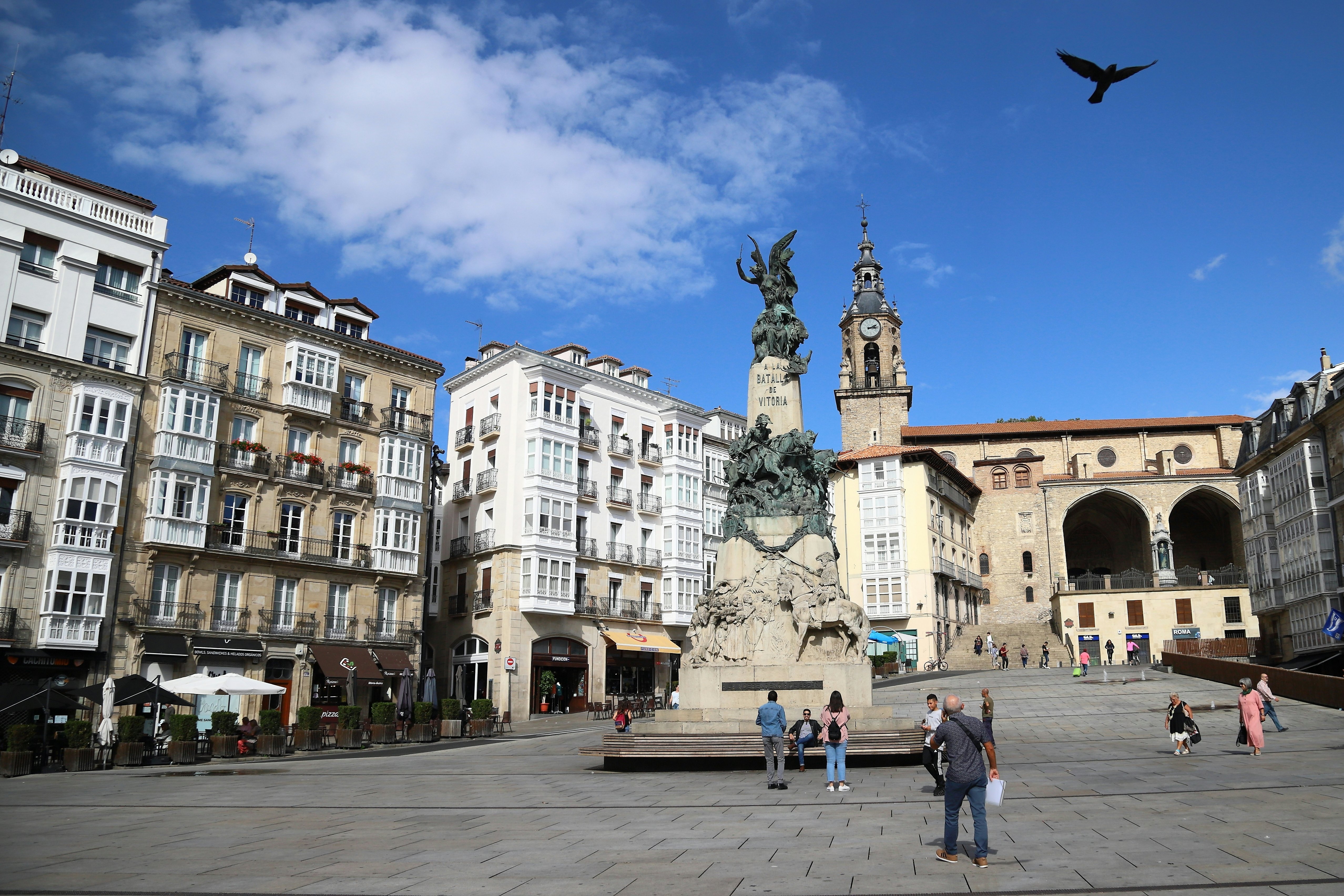 People are seen in a city square, with a large sculpture-topped monument in the center and historic buildings along the perimeter.