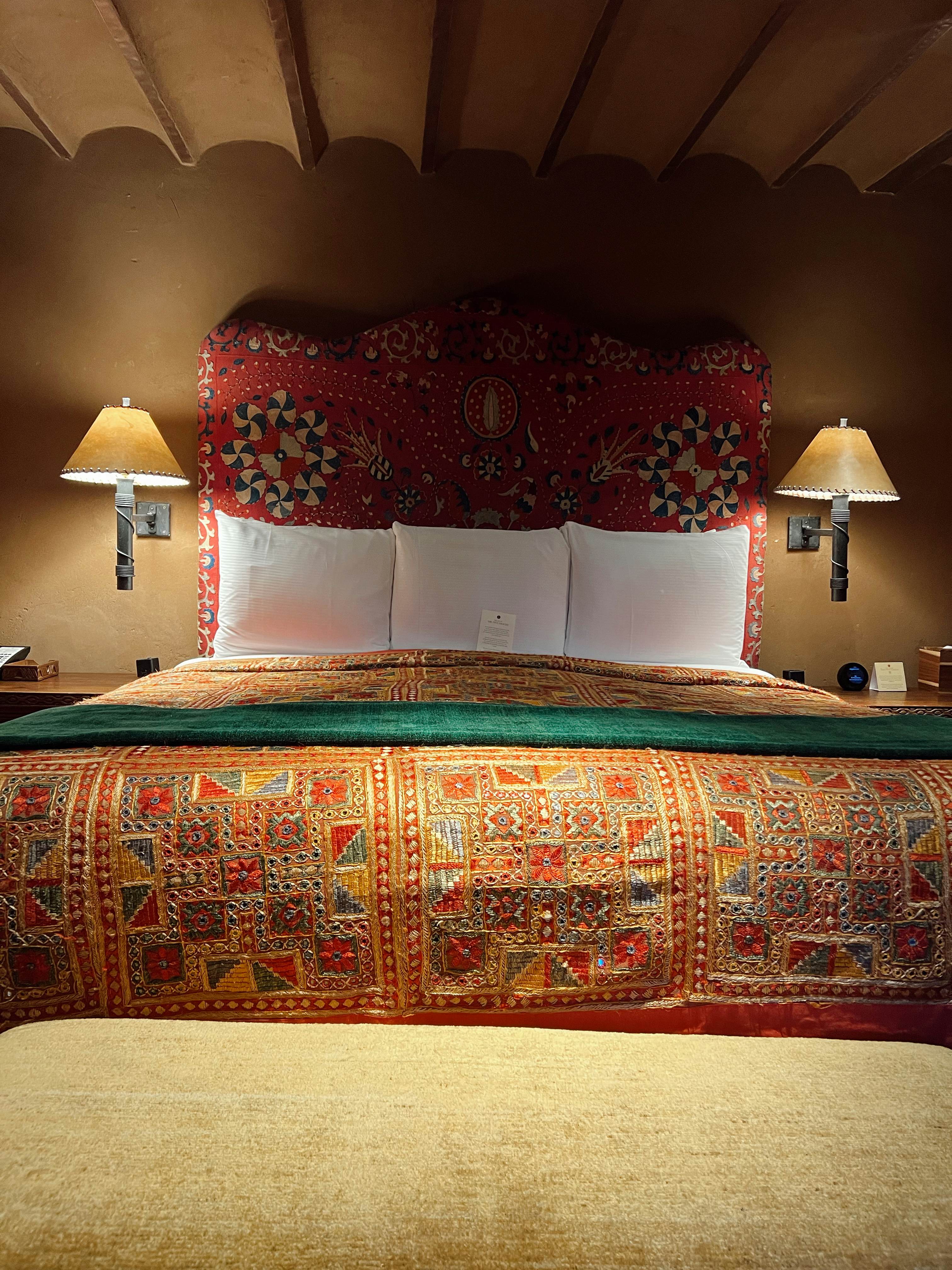 A bed with a patterned spread, three white pillows and lit sconces on either side.