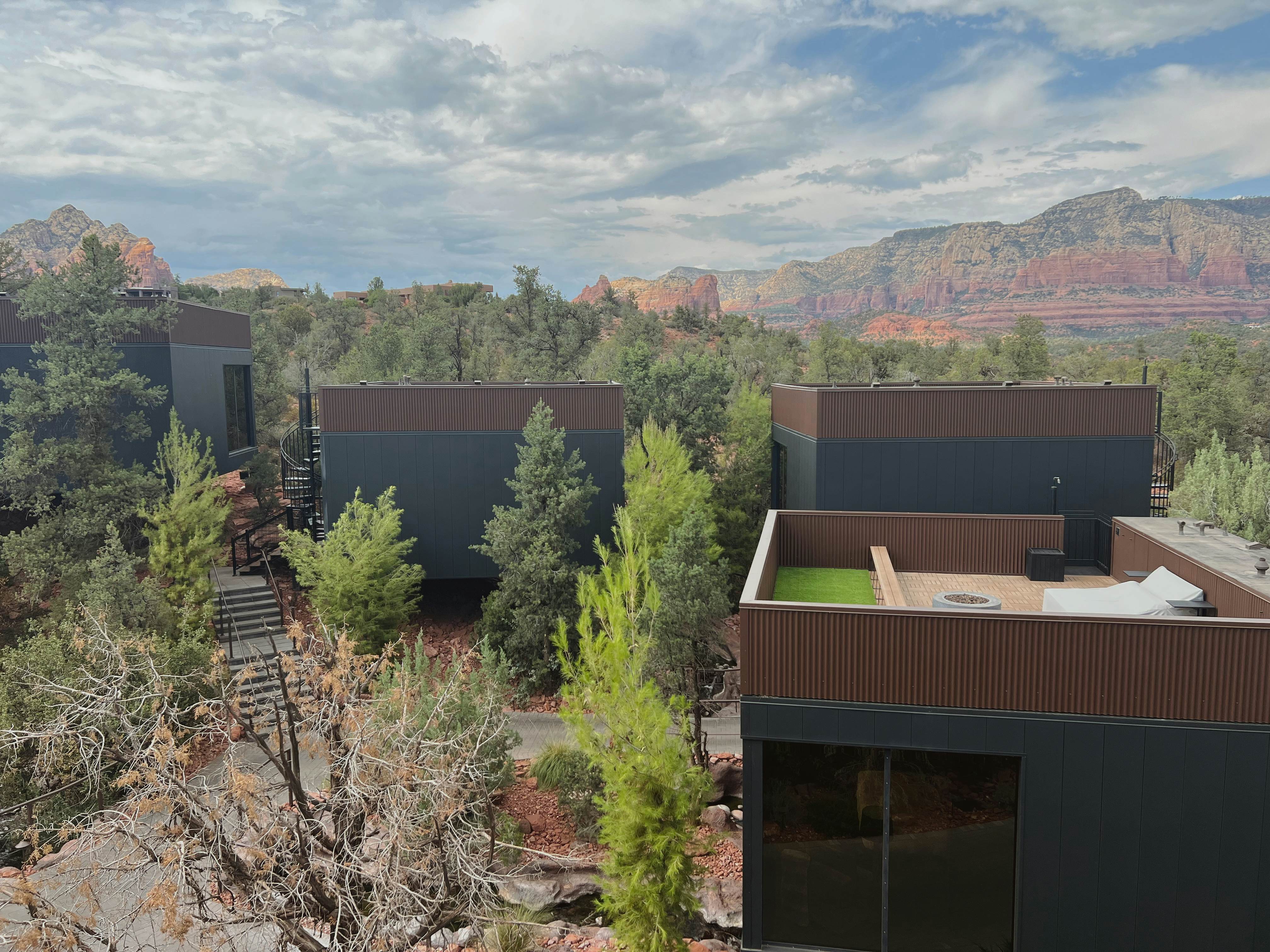 Structures surrounded by trees with reddish rock formations in the background under cloudy skies.