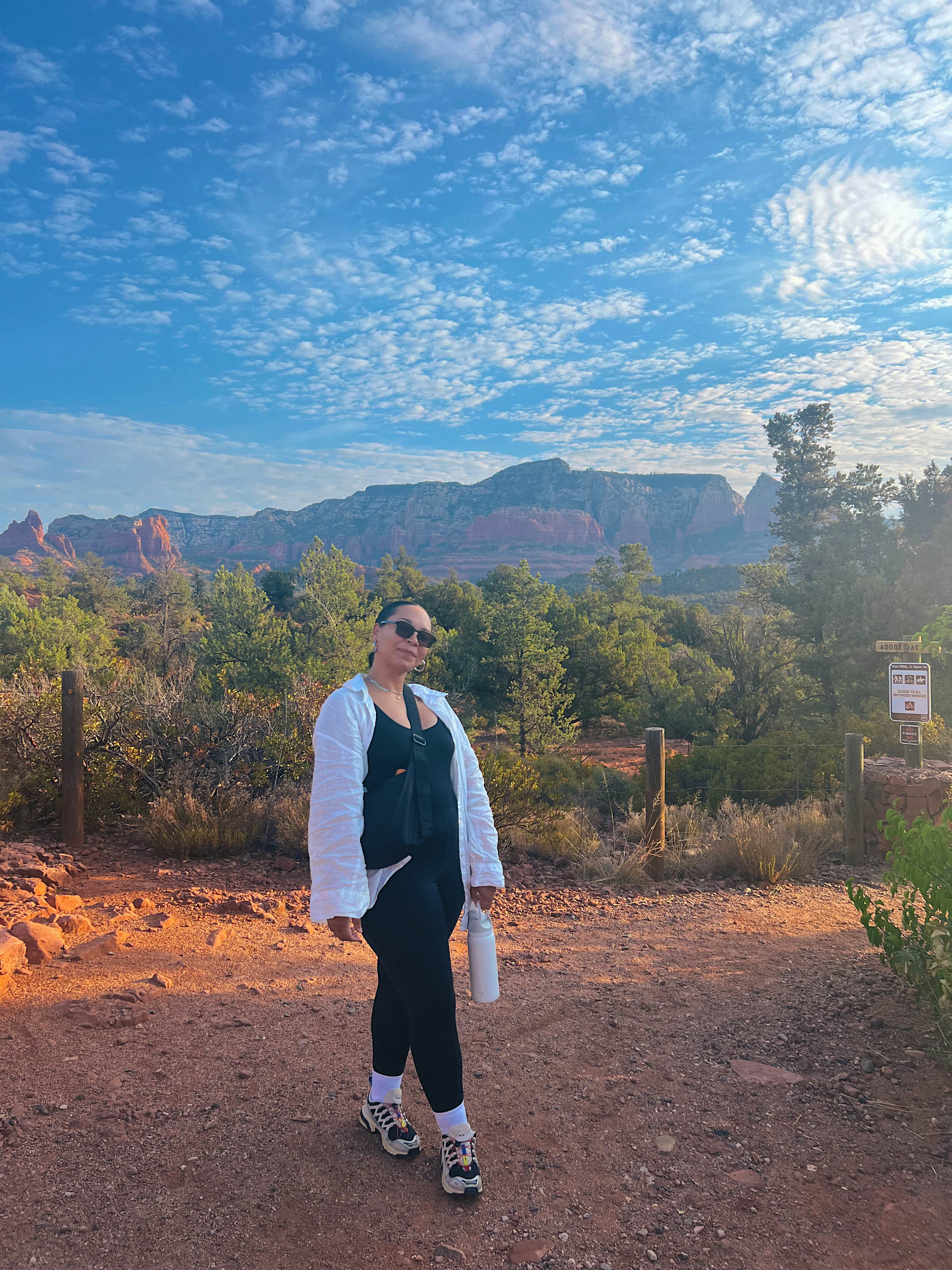 A person stands on a trail with trees and reddish rock formations in the background.