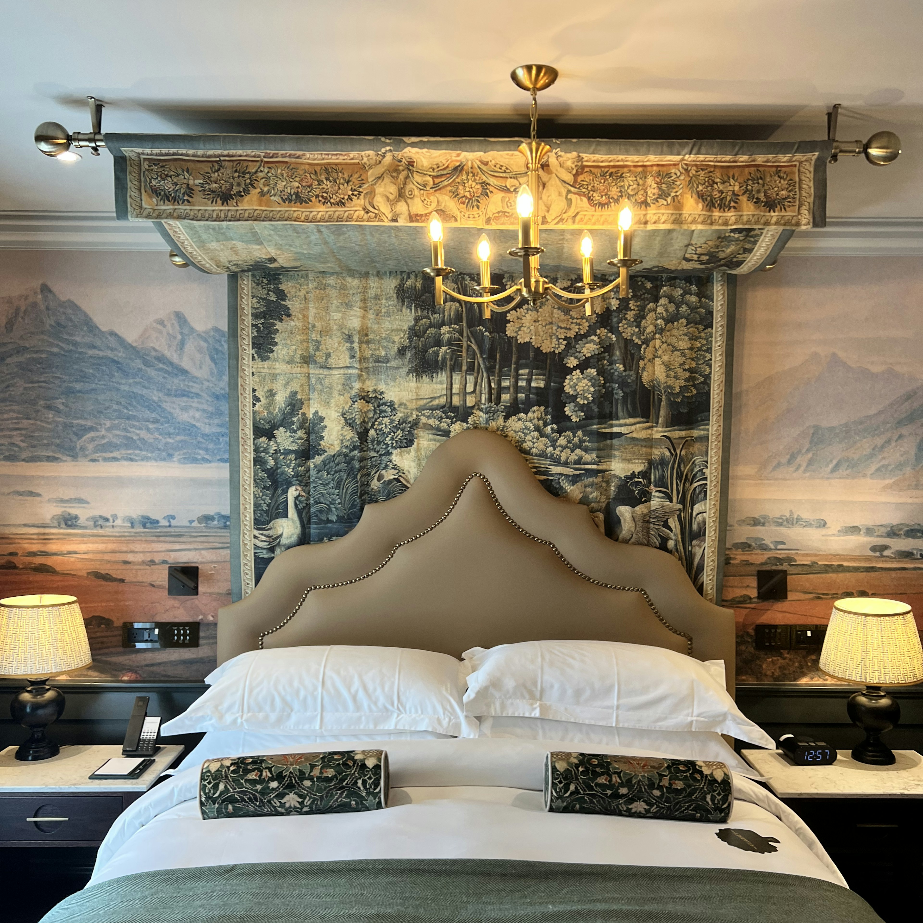 A bed with a white covering and round patterned pillows; the triangular headboard is beige, and the wall behind it has layered pieces of artwork.