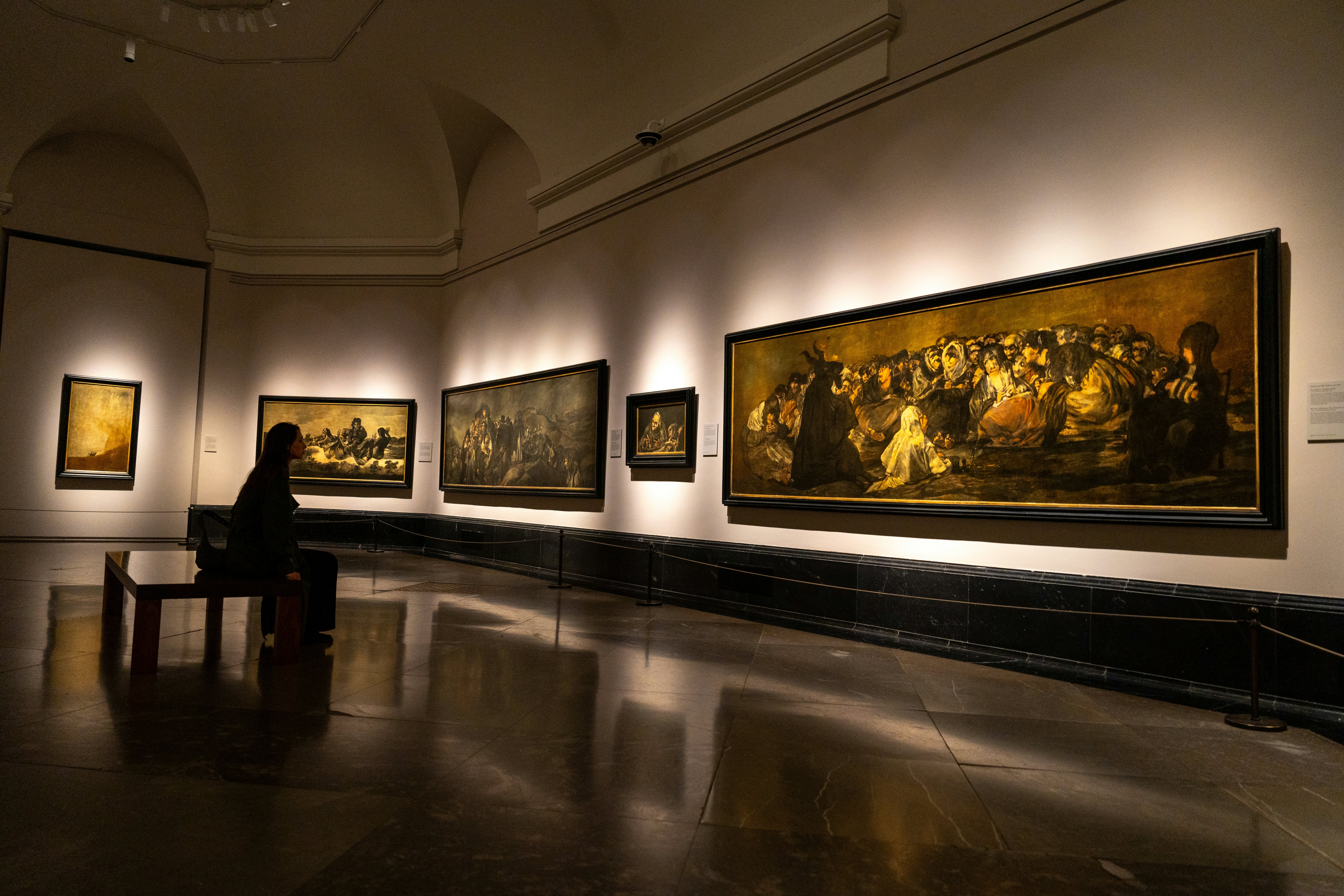 A person sits on a bench in a darkened gallery looking at large paintings on a wall.