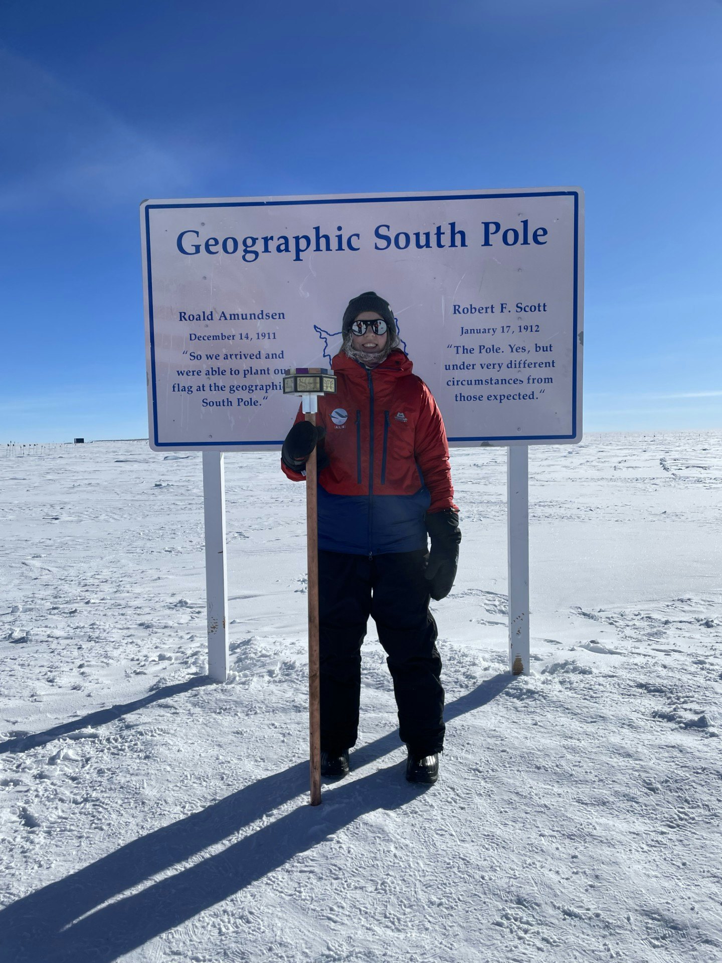 A woman stands in front of a sign in a snow field with the words "Geographic South Pole" on it.