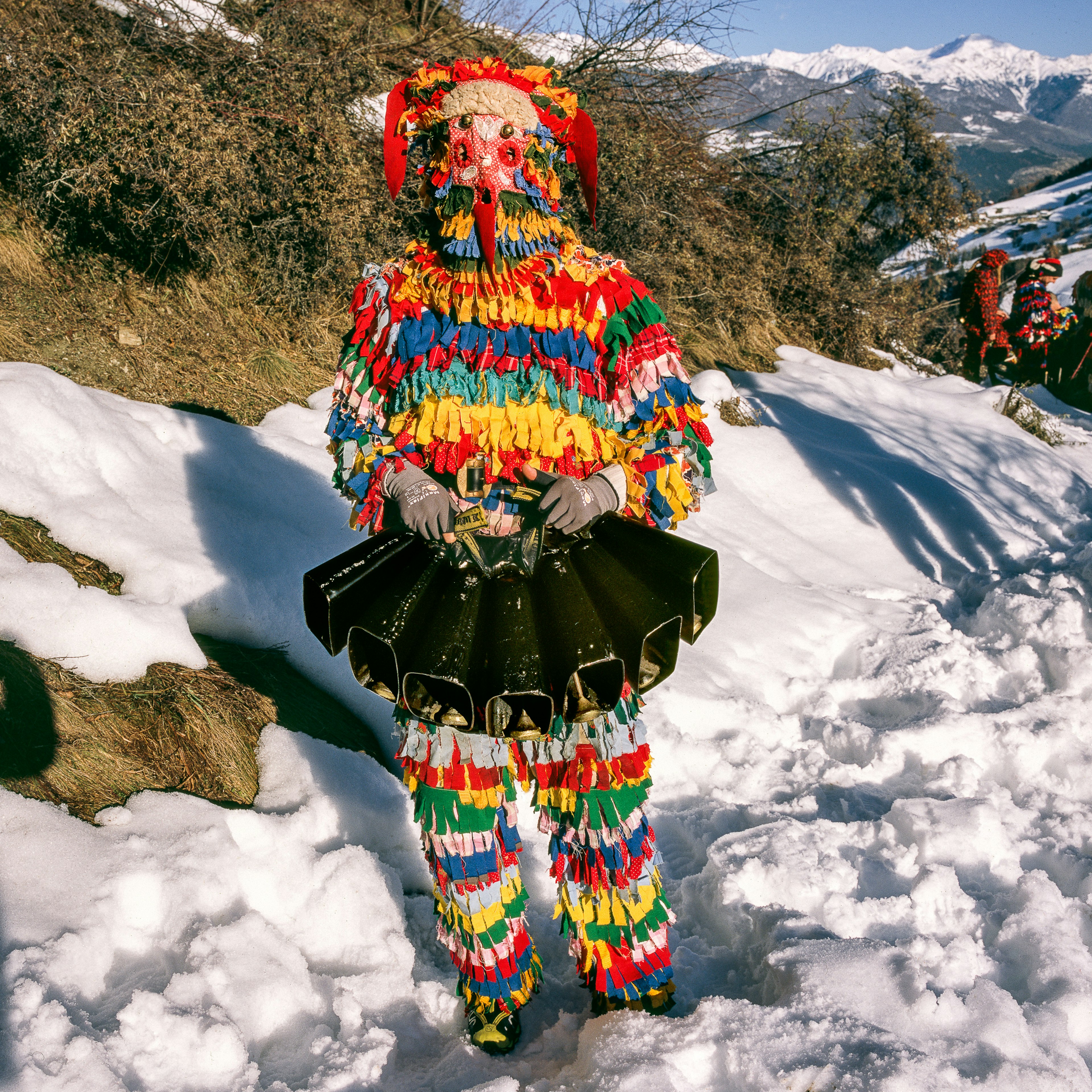 A person in a multicolored costume stands in the snow holding a black object.