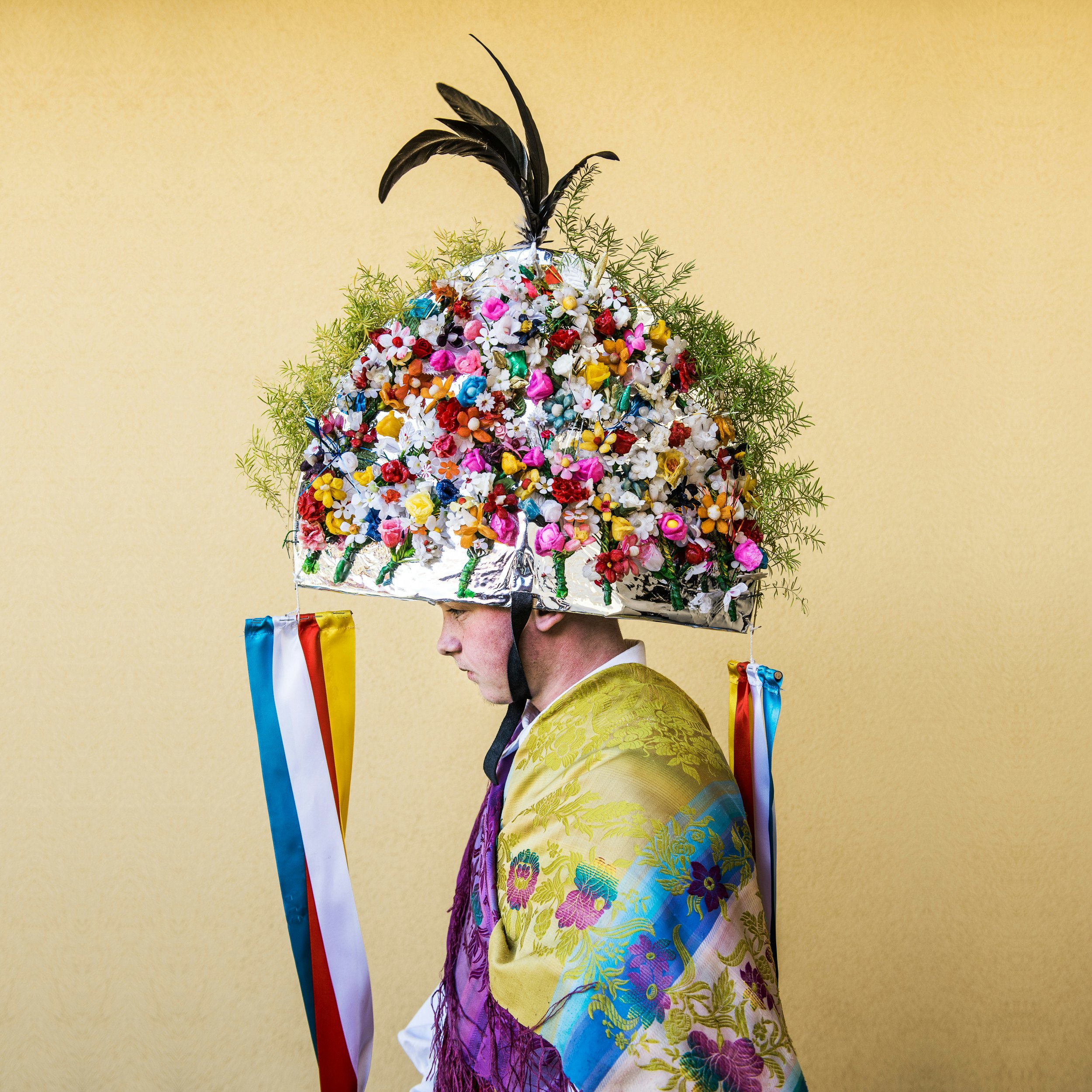 A person wears a large round headpiece covered with flowers and a patterned costume.