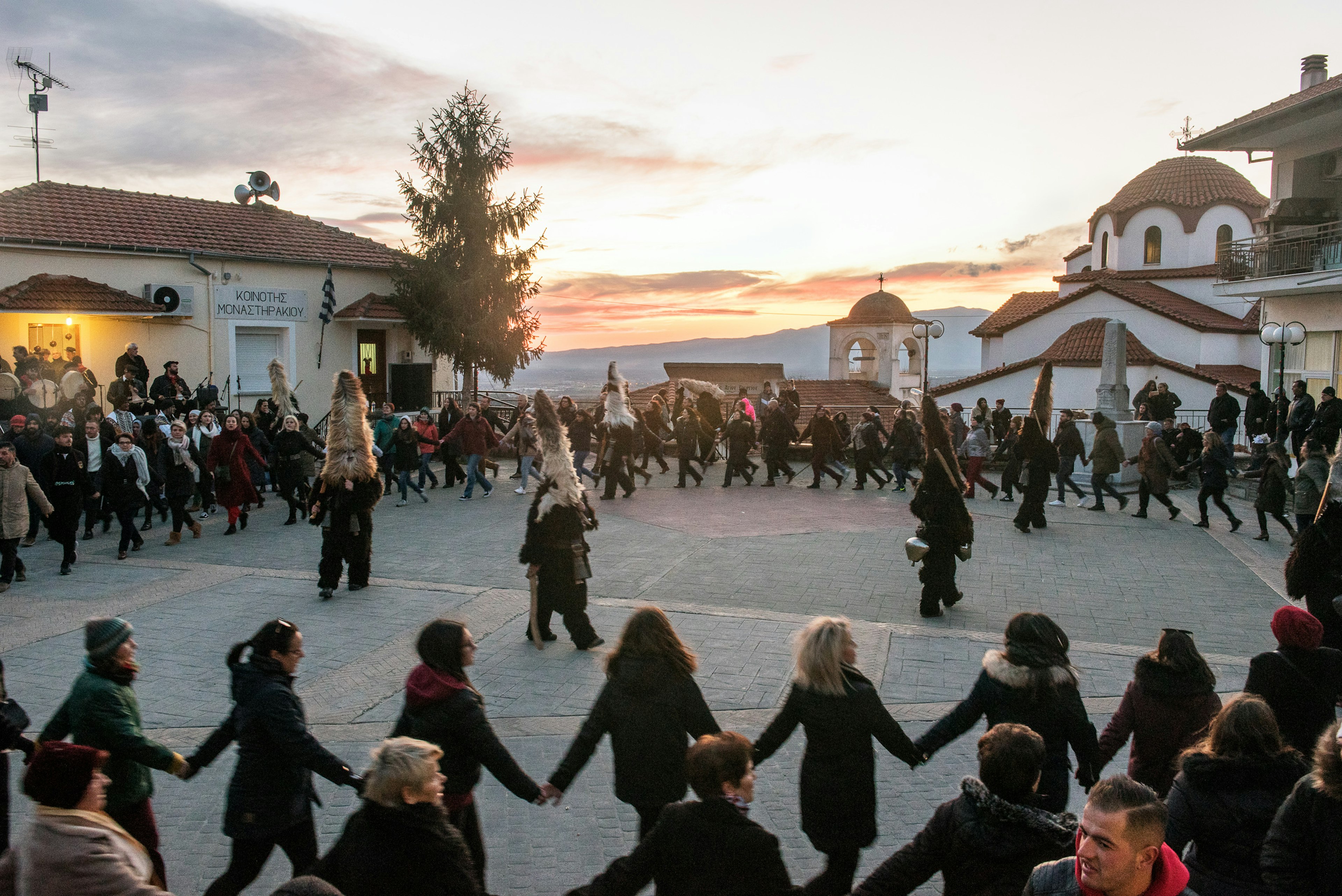 Dancers join hands and circle around an open plaza at sunset. Several costumed figures are in the middle of the circle.