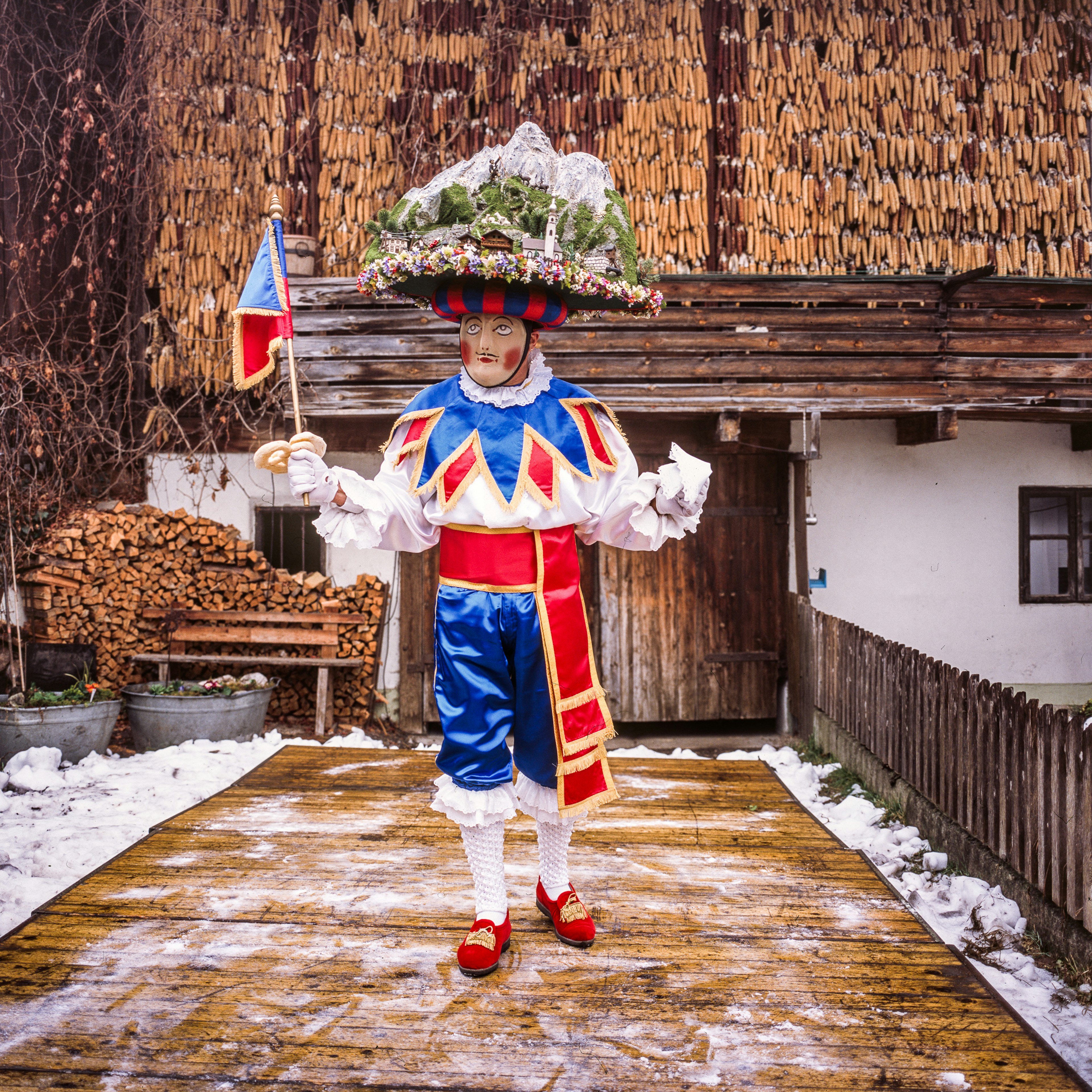 A person wearing a red, white and blue costume wears a mask and carries a flag in front of a traditional home.