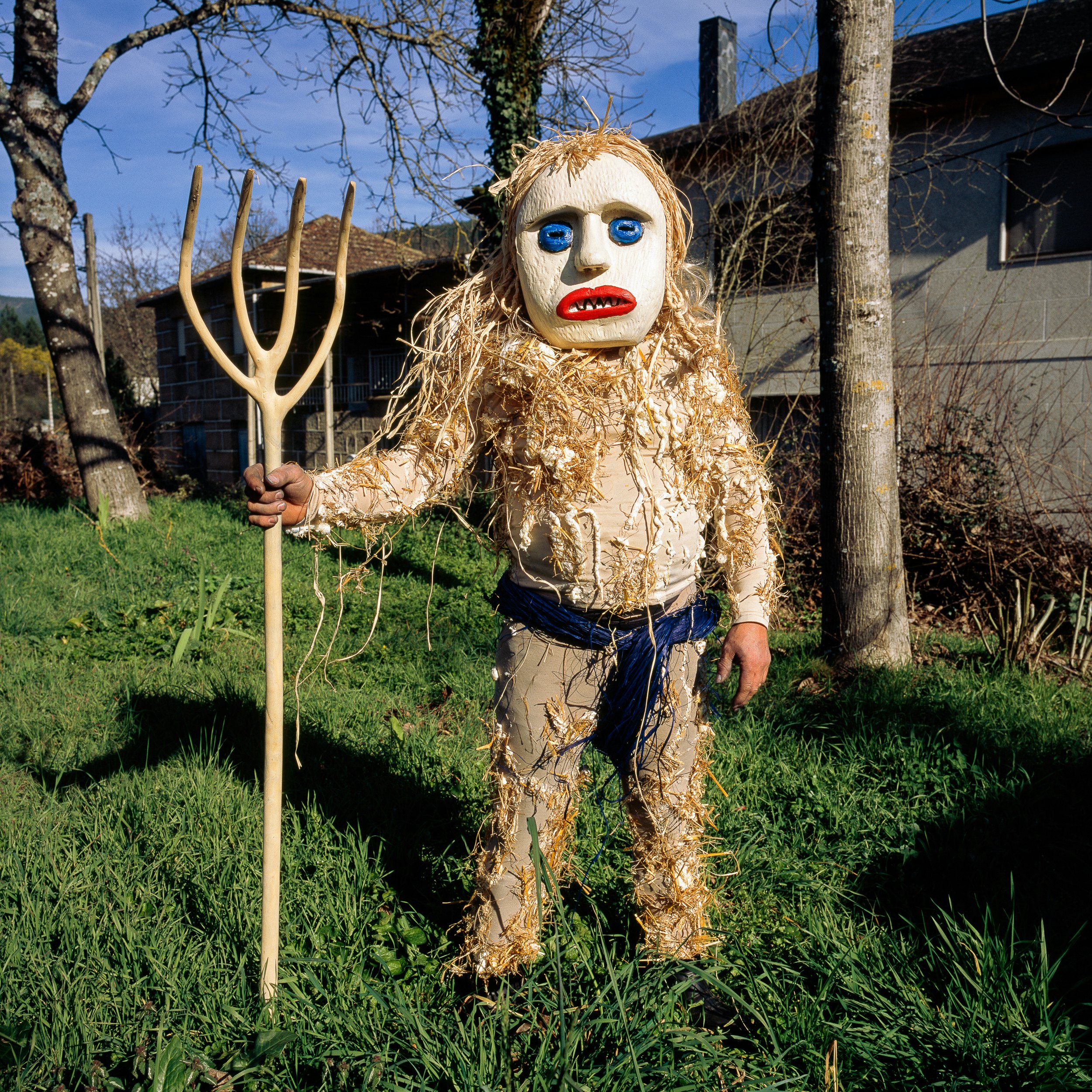A masked person wearing a costume holds a pitchfork in the grass.