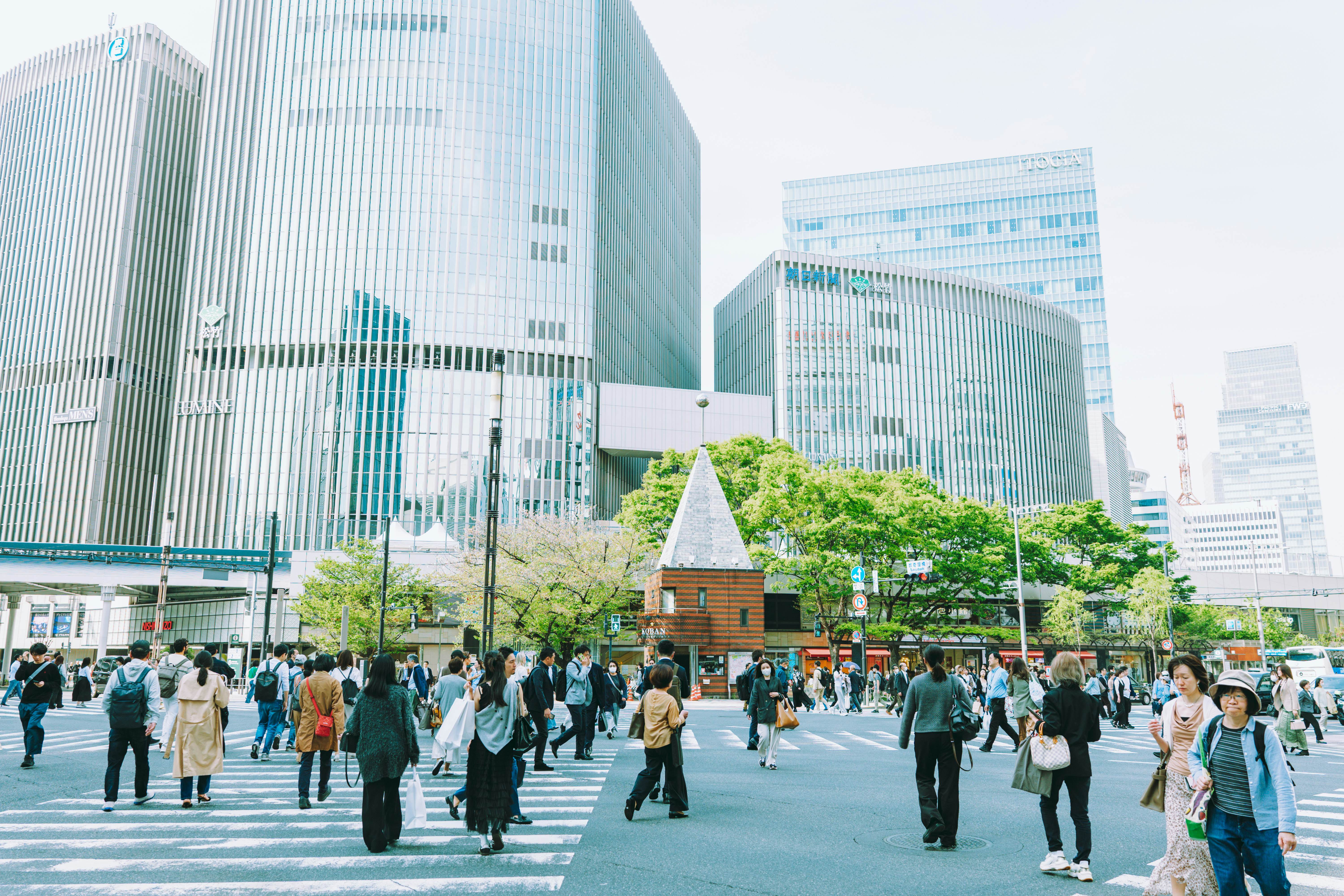 People cross a large intersection with glass skyscrapers on the other side.