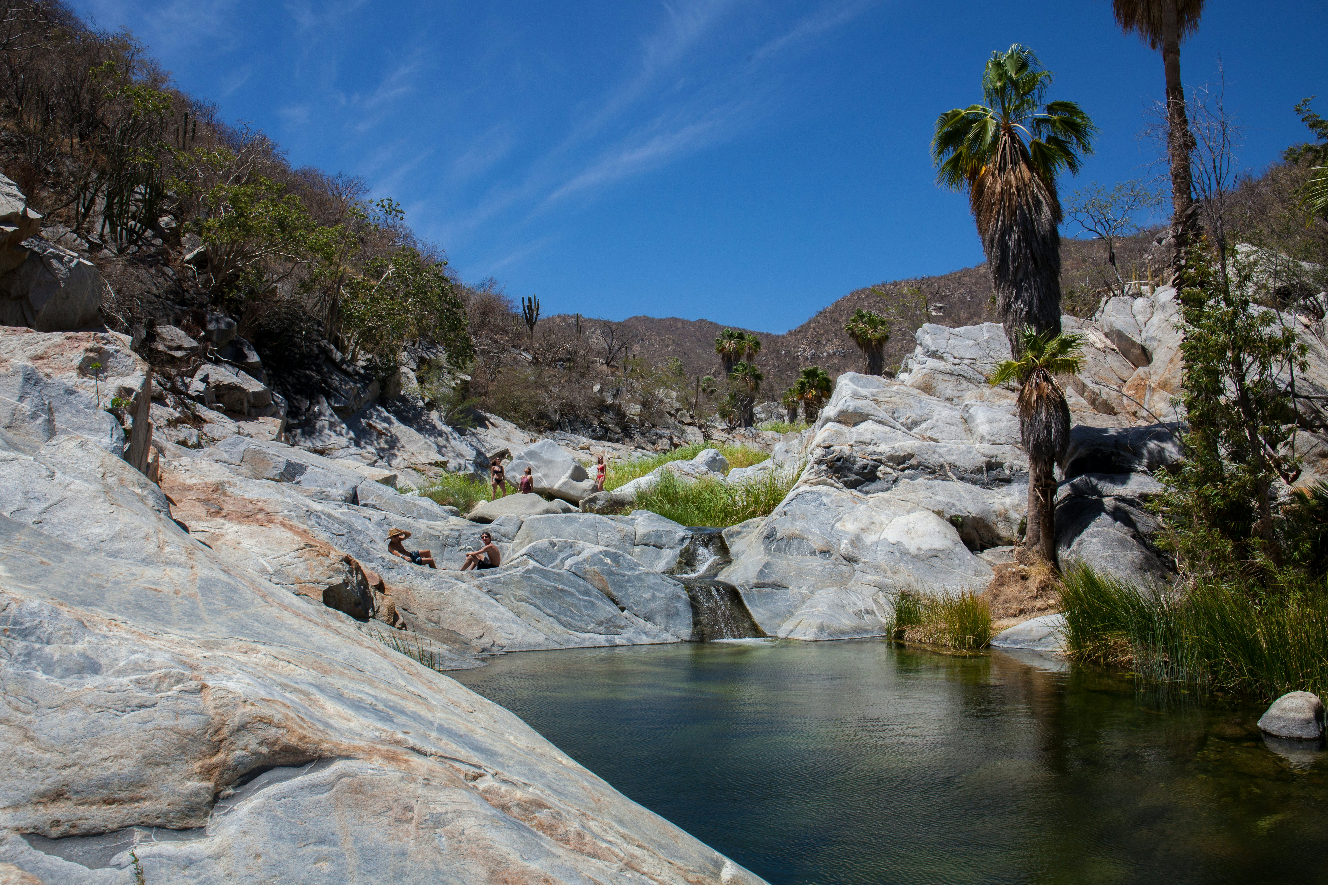 People sit on large gray rocks in sunshine by a dark pool of water; dry mountains and vegetation are in the background.